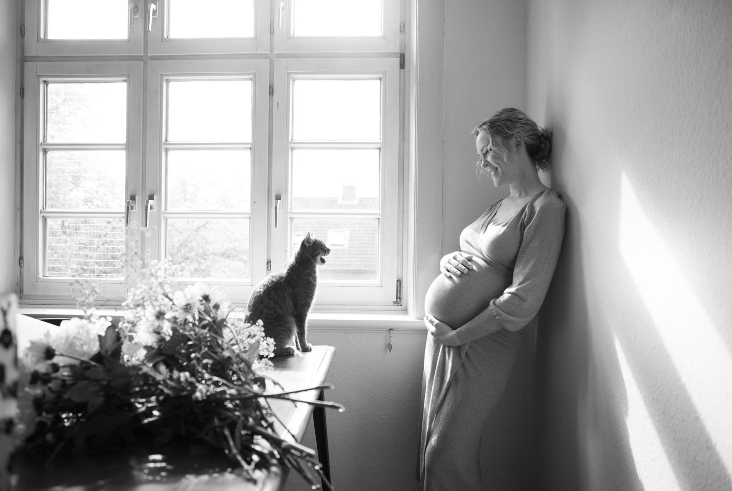 Black cat perches on windowsill beside flowering plants in monochrome photograph.