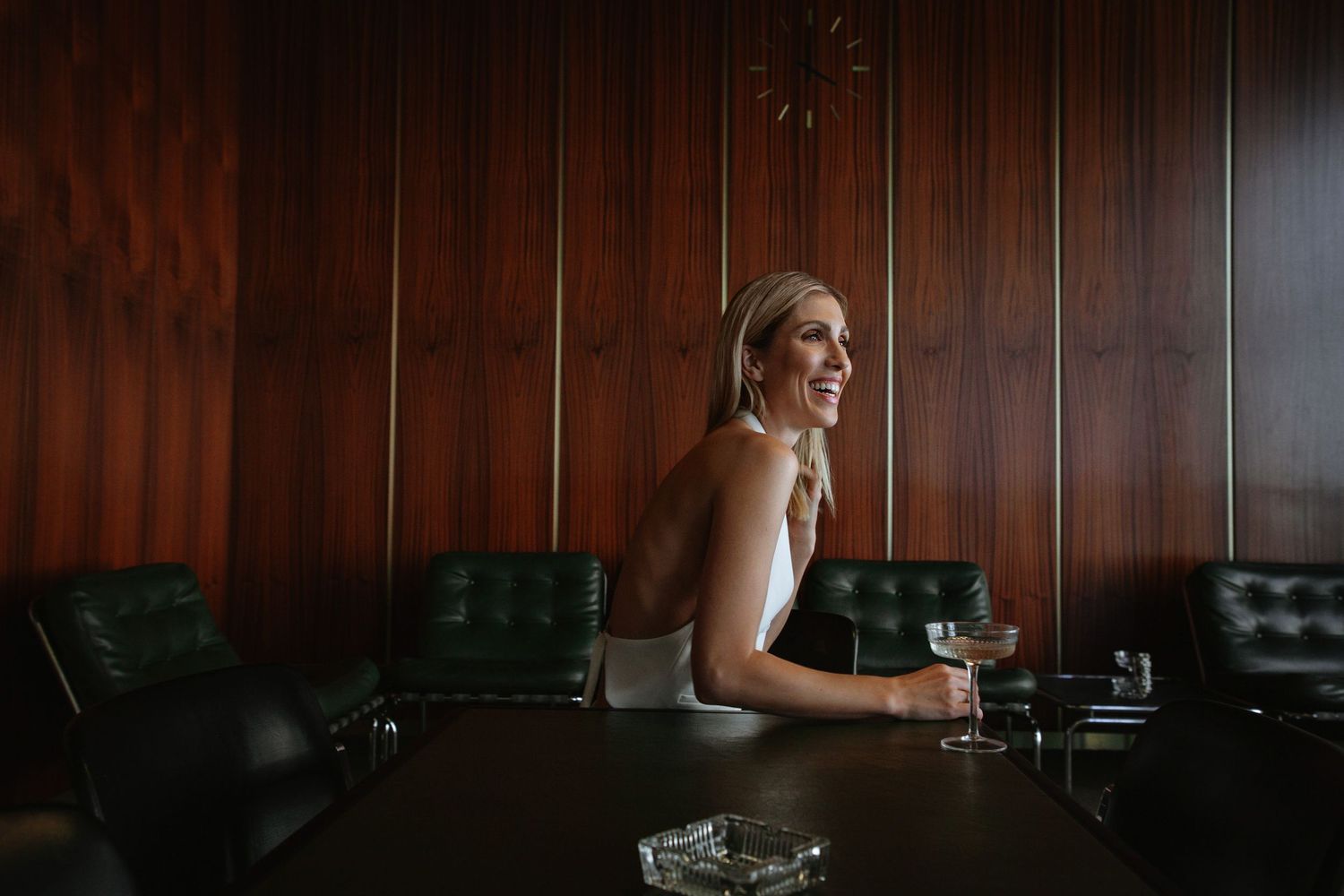 Woman in evening attire seated at dark bar with wine glass and mood lighting.