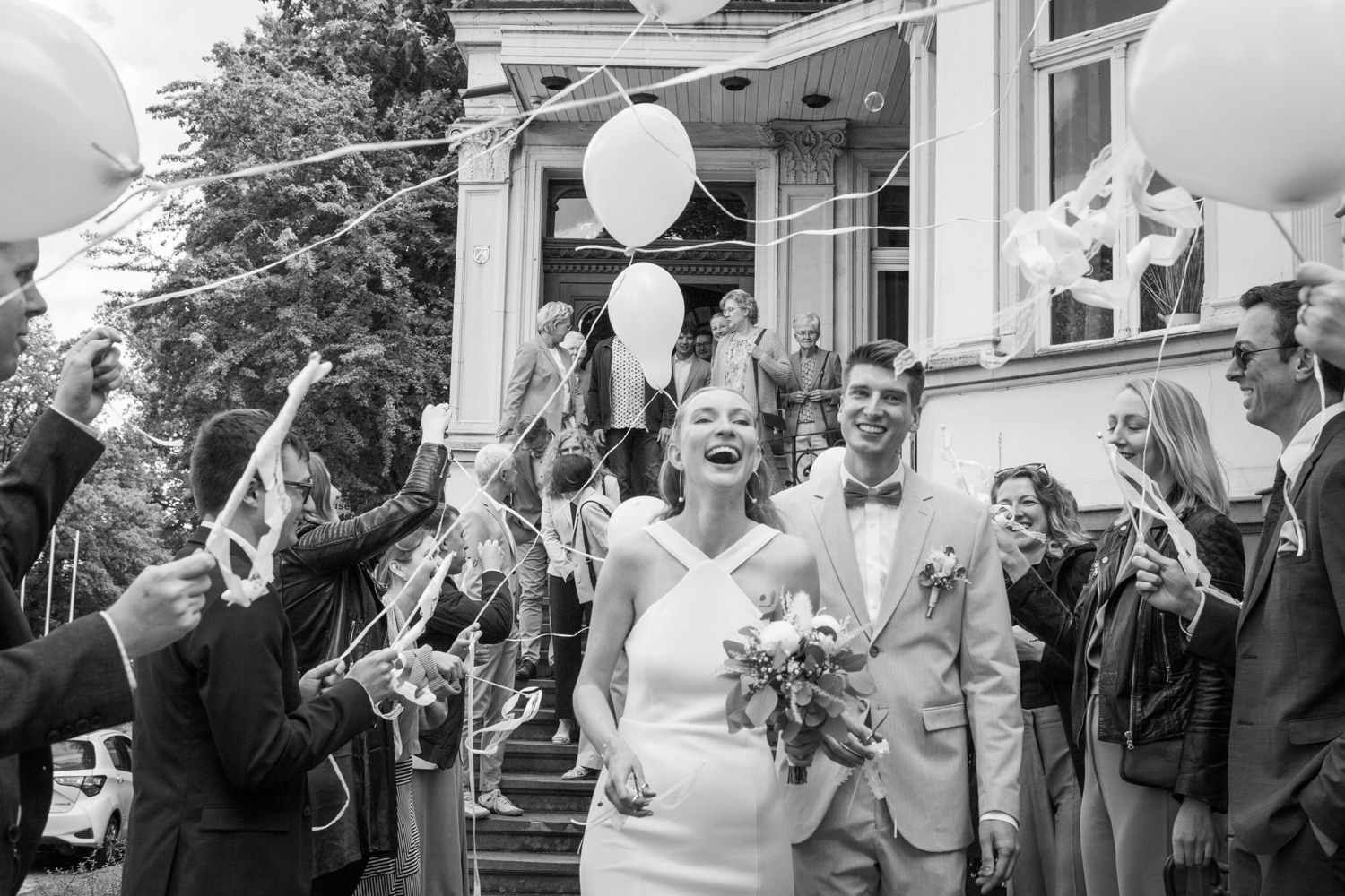 Joyful wedding celebration outside with guests throwing confetti and holding balloons in black and white photography.