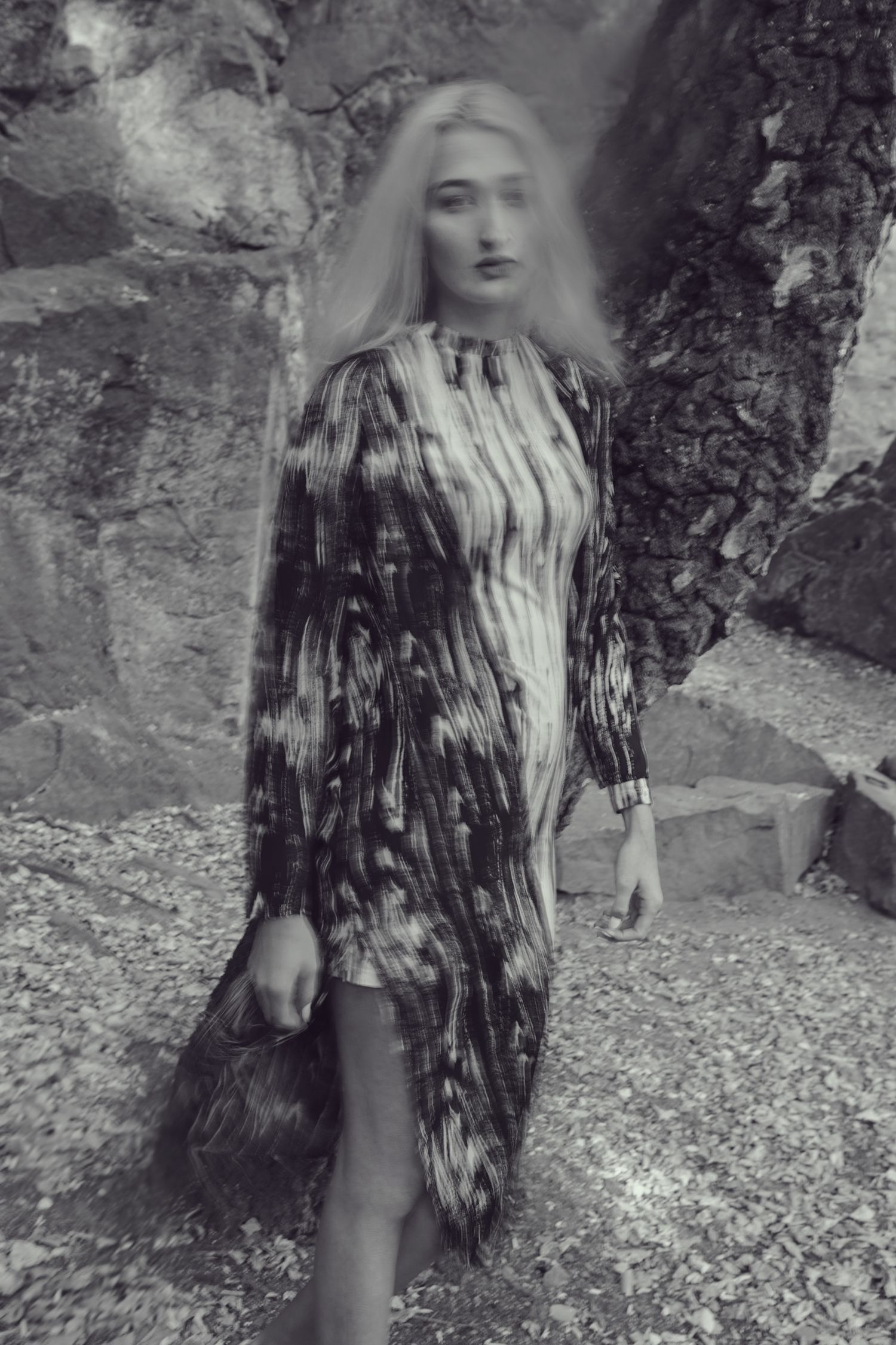 Black and white photo of a person in a patterned dress standing against rocky wall.