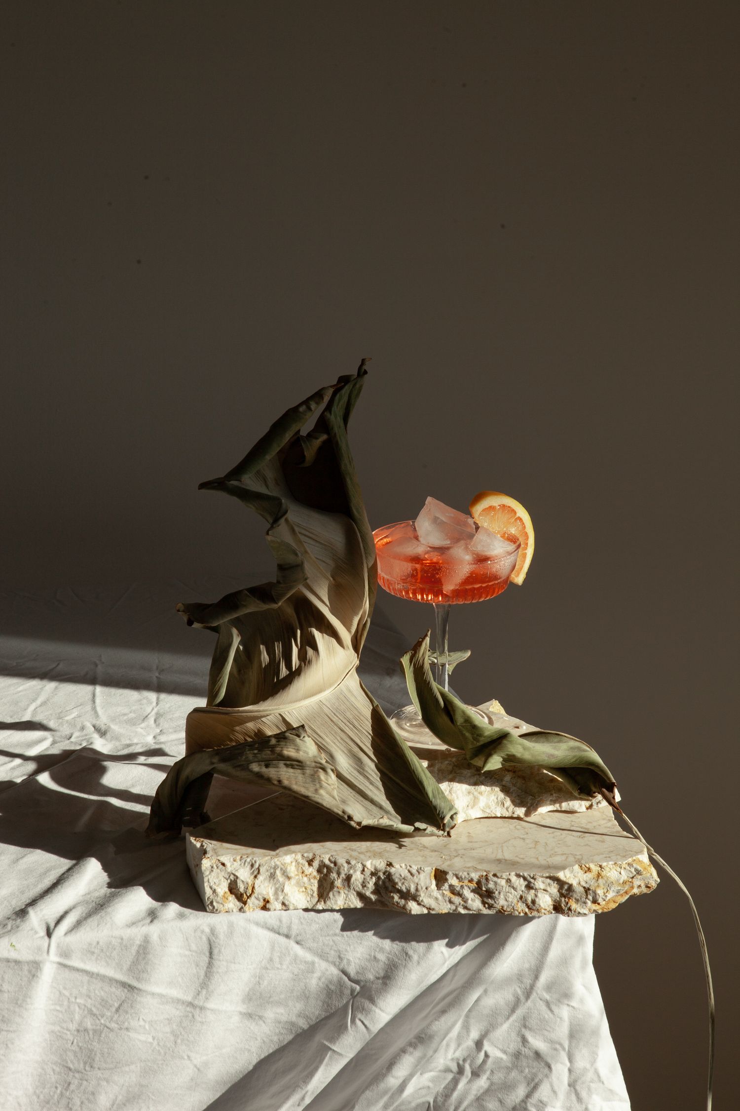Moody still life with a pink cocktail, lemon garnish and monstera leaves against dark background on stone surface.
