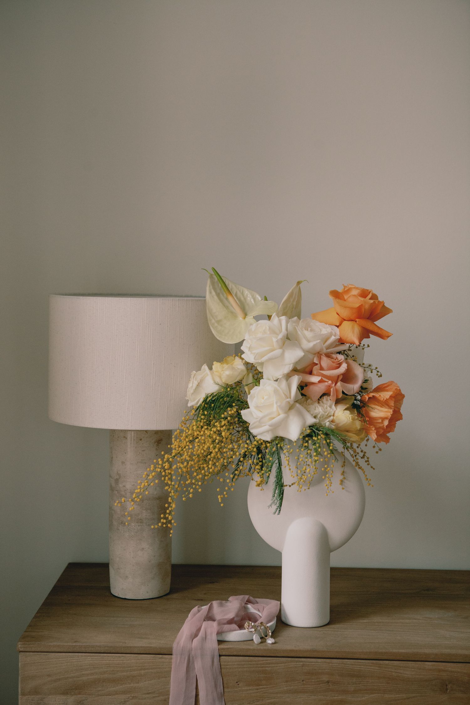 Modern floral arrangement with roses and mimosa in white vase next to concrete table lamp.
