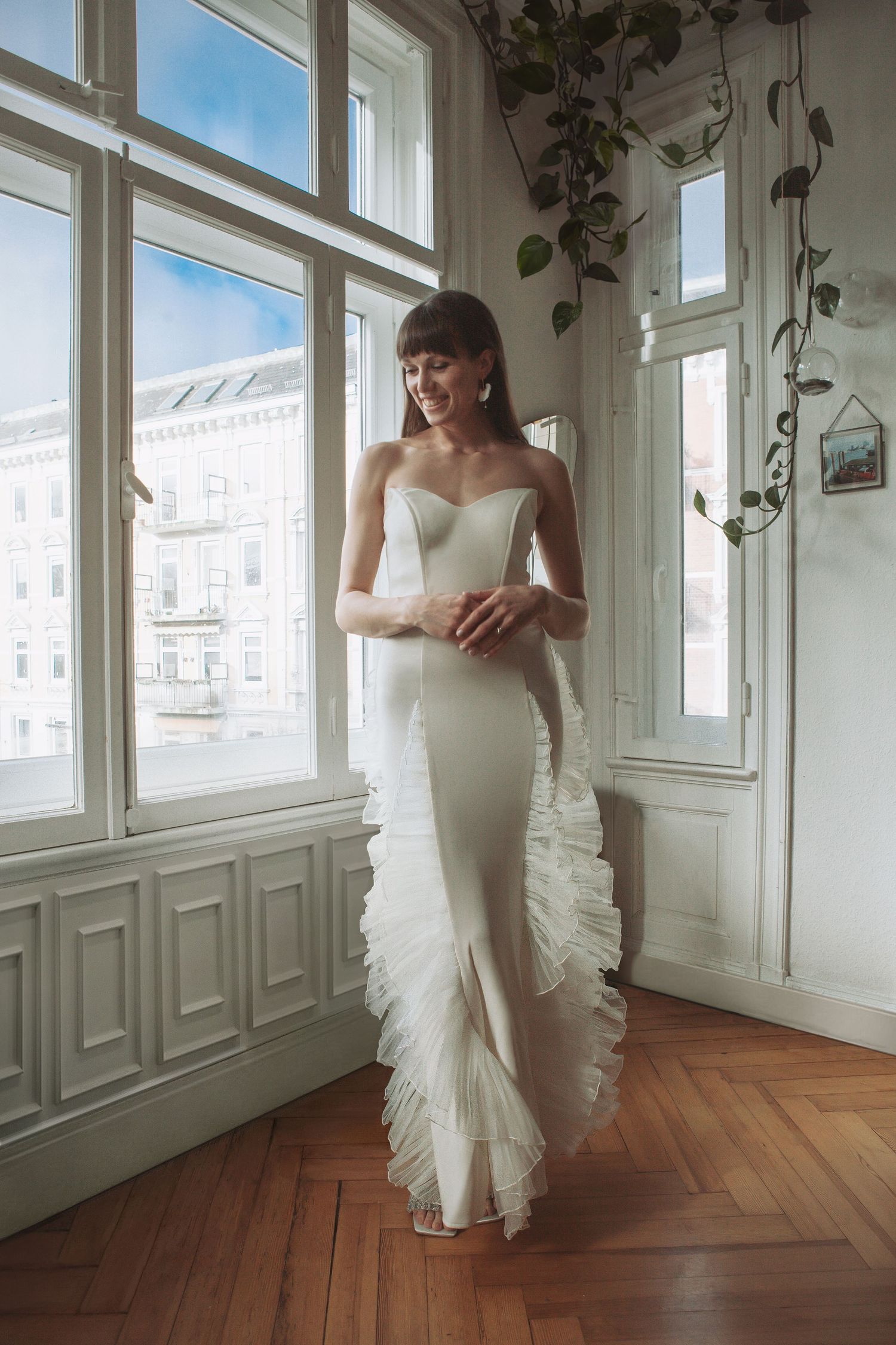 Bride in form-fitting white gown with ruffled train poses by sunny apartment windows.