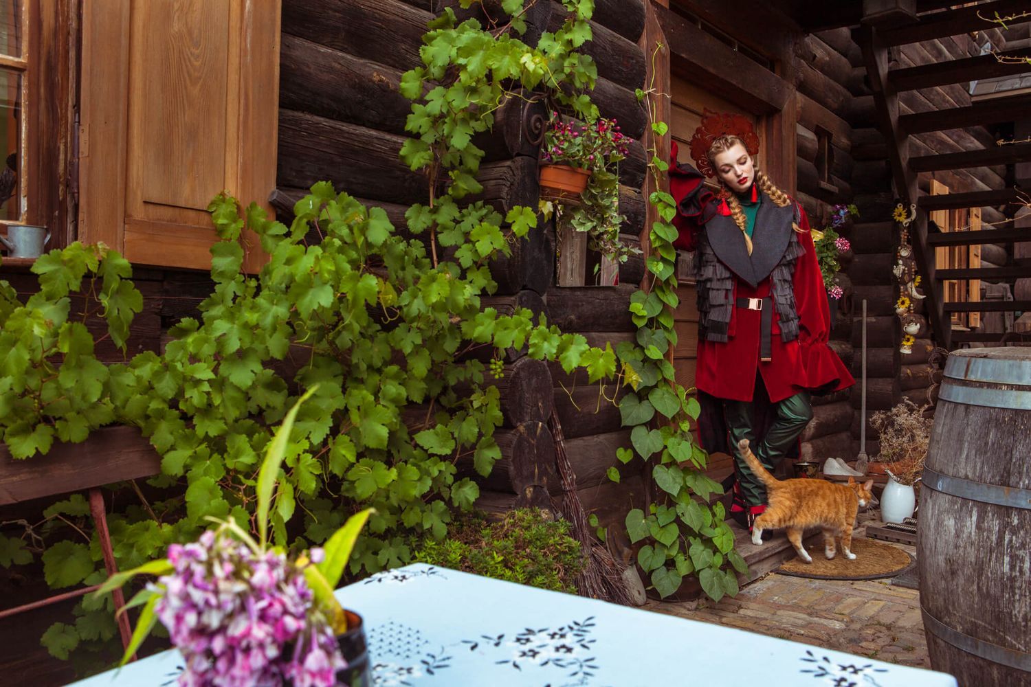 Rustic countryside fashion scene with red coat by wooden cabin with cat and garden elements.