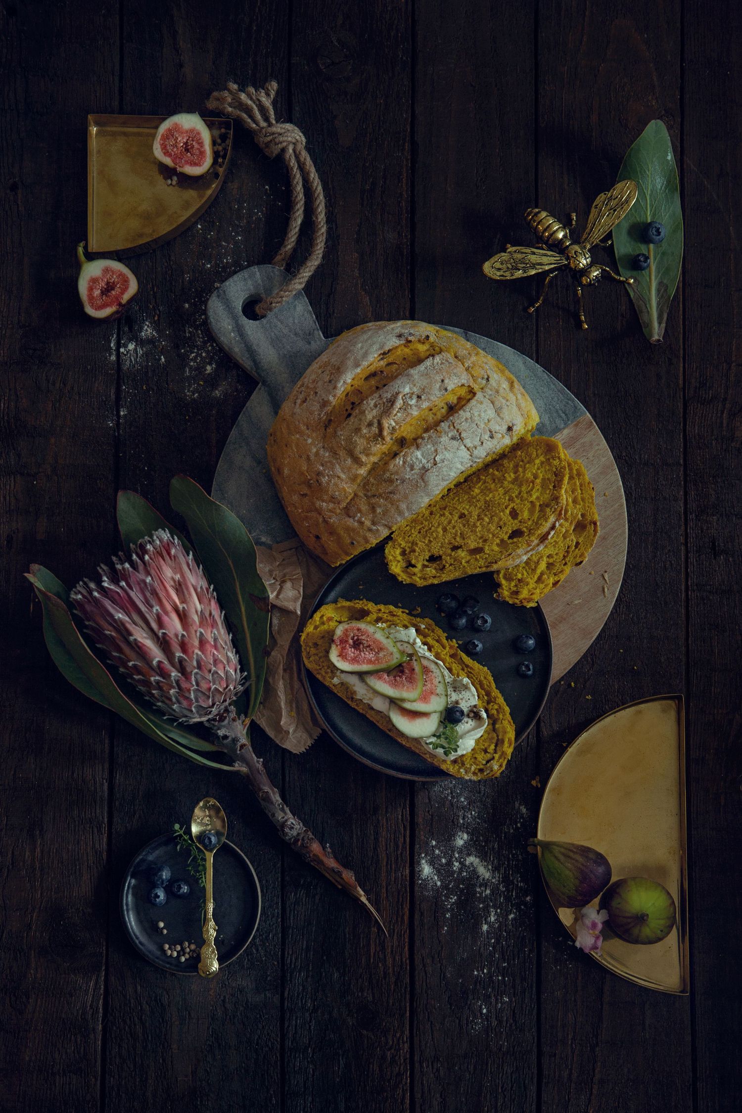 Rustic food styling with bread, figs, and protea flowers on dark wooden surface.