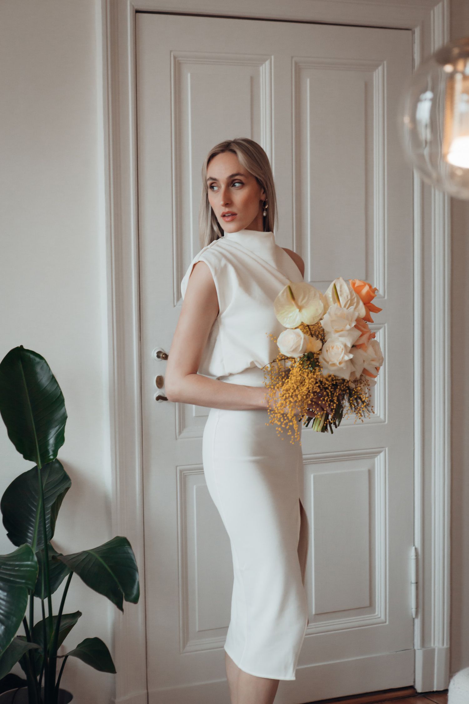 Elegant bride in white fitted dress holding a bouquet of yellow and cream flowers by a paneled door.