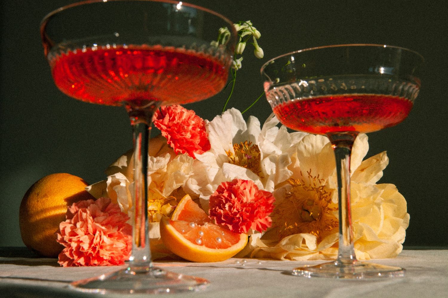 Two red cocktails in coupe glasses surrounded by flowers and citrus garnishes.