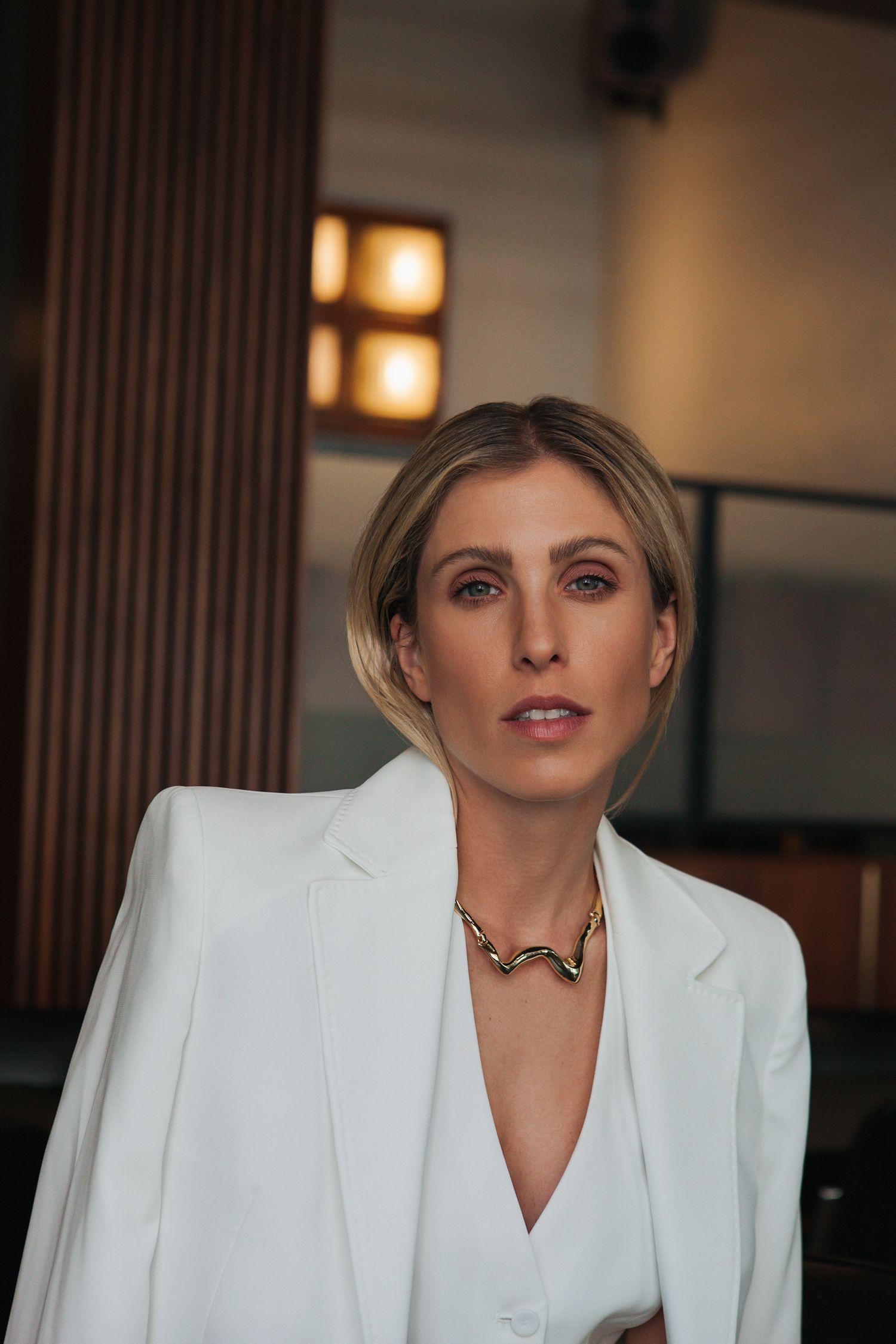 Elegant portrait of woman wearing white blazer and gold chain necklace against warm interior lighting.