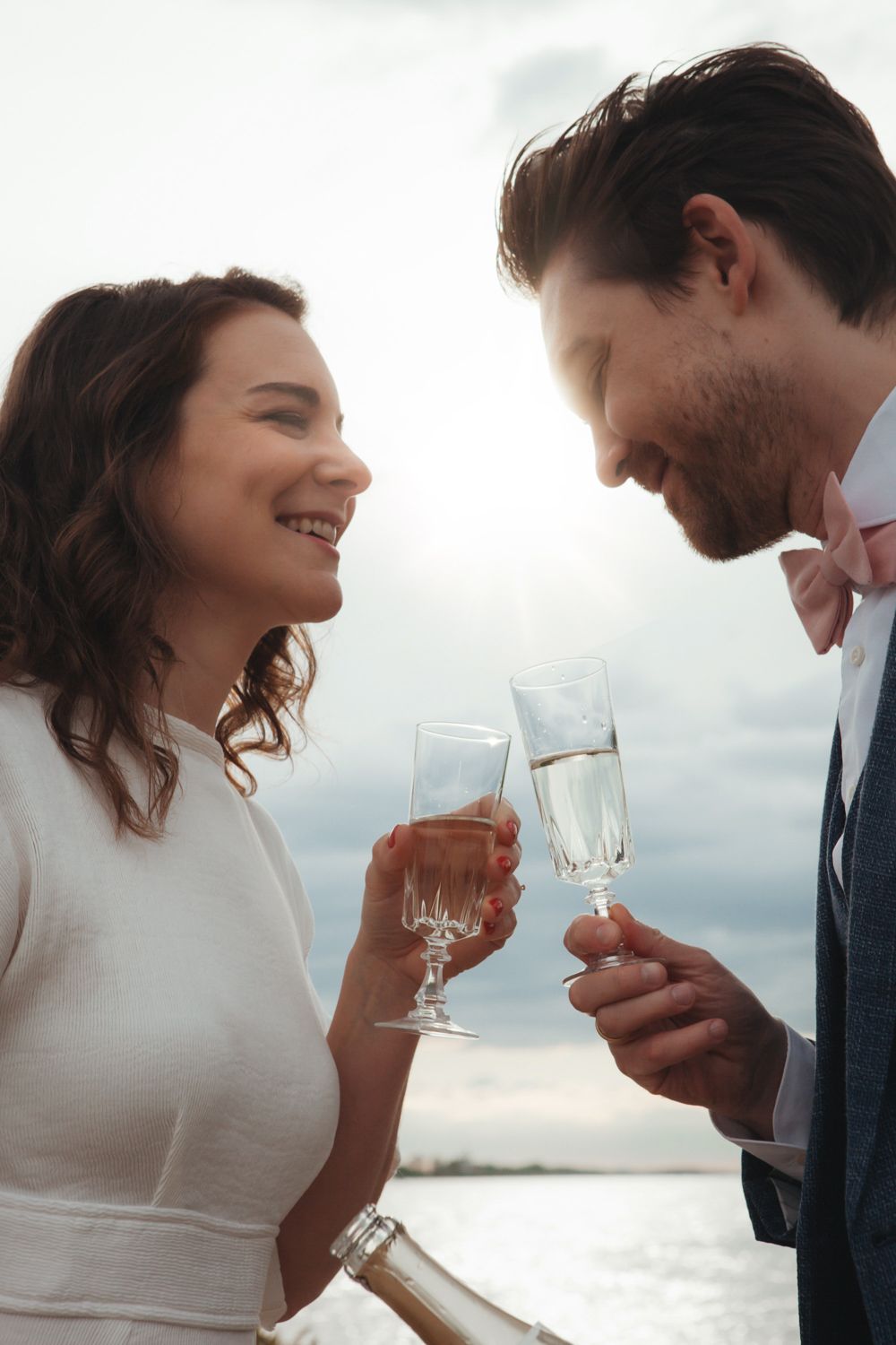 A couple shares an intimate moment together holding wine glasses against a bright sky backdrop.