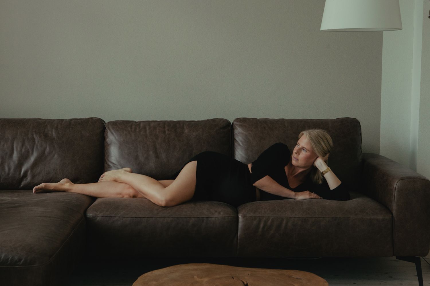 Someone relaxes on a dark brown leather couch in a dimly lit living room, stretching out across the cushions.