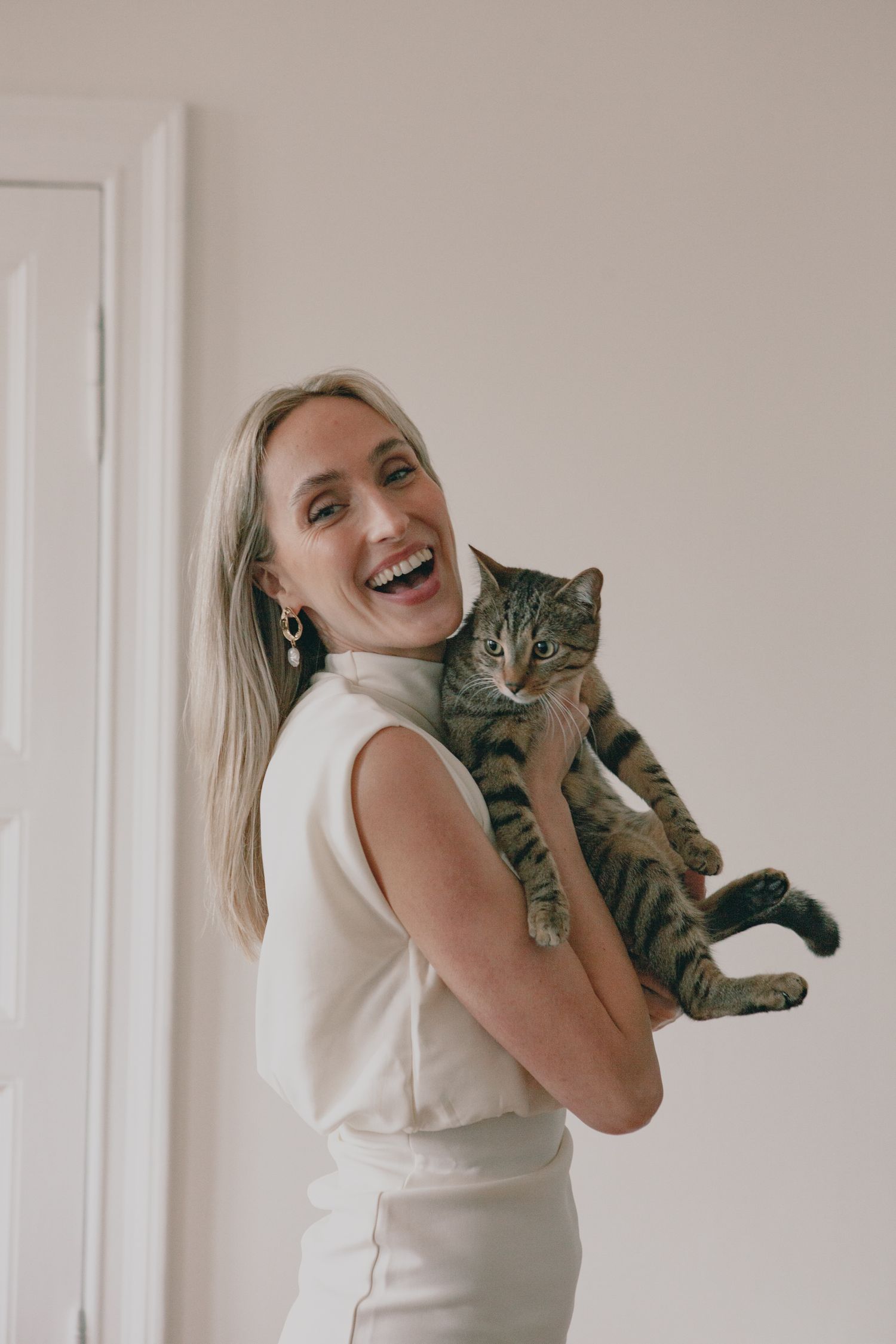 Woman in white outfit holding striped tabby cat while laughing against neutral wall.