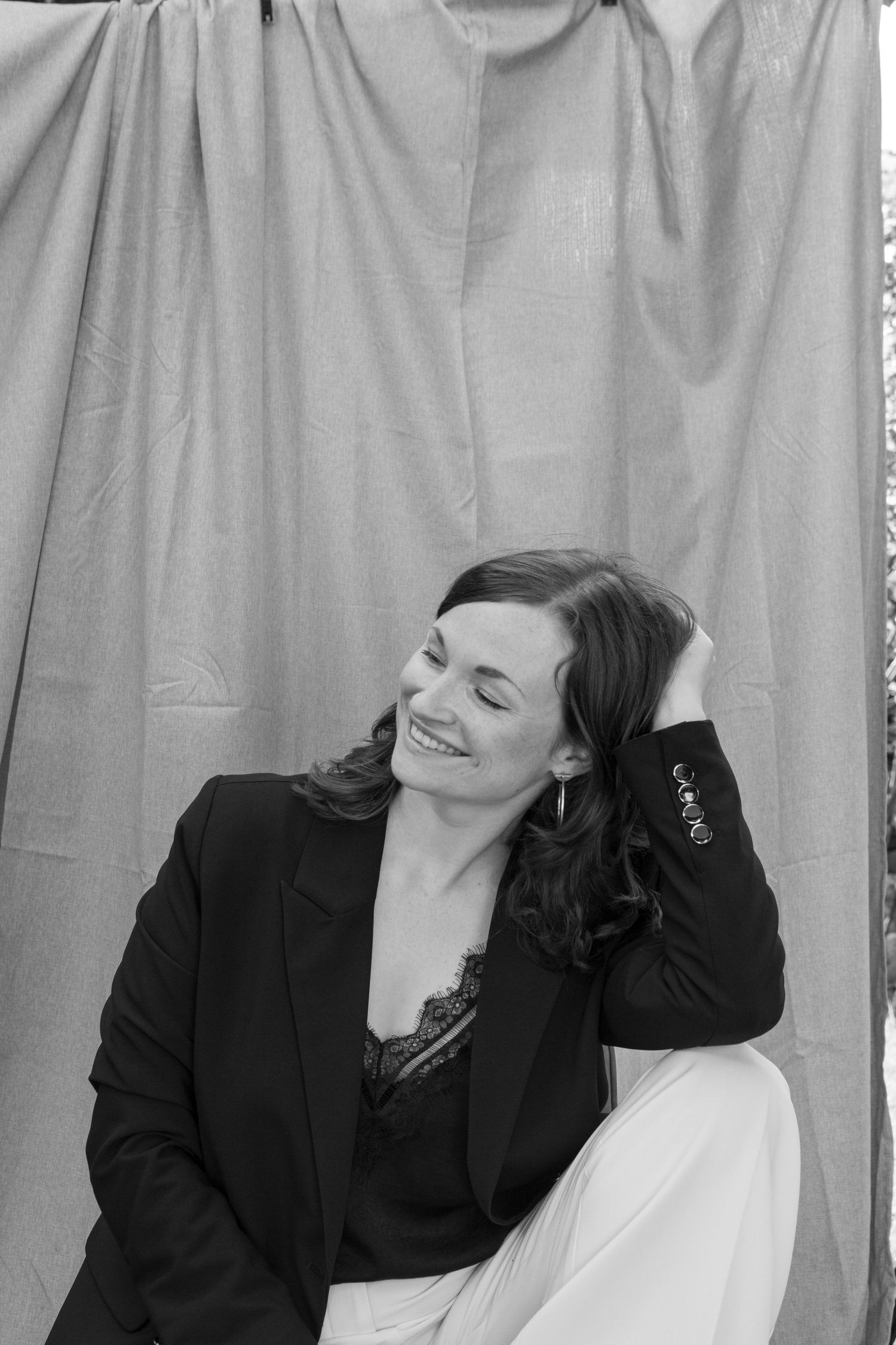 A black and white portrait of a person in a black blazer and white pants relaxing with a joyful smile.