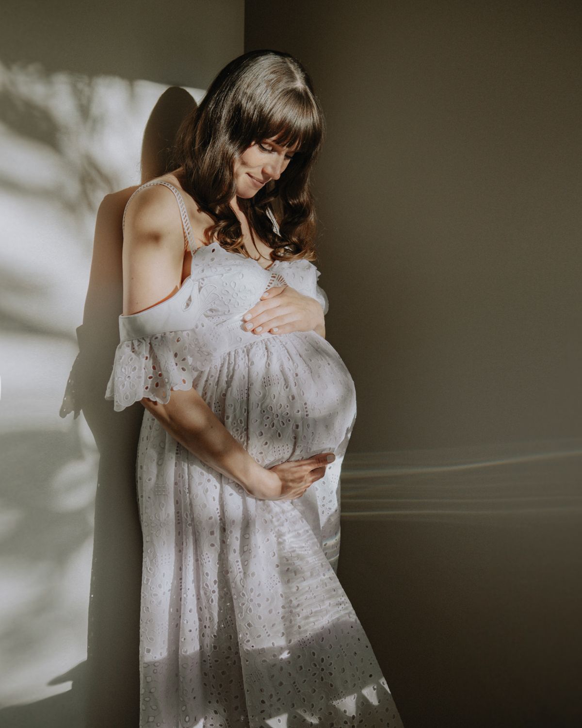 Expectant woman in a flowing white lace dress stands against a wall with dramatic shadows falling across her form.