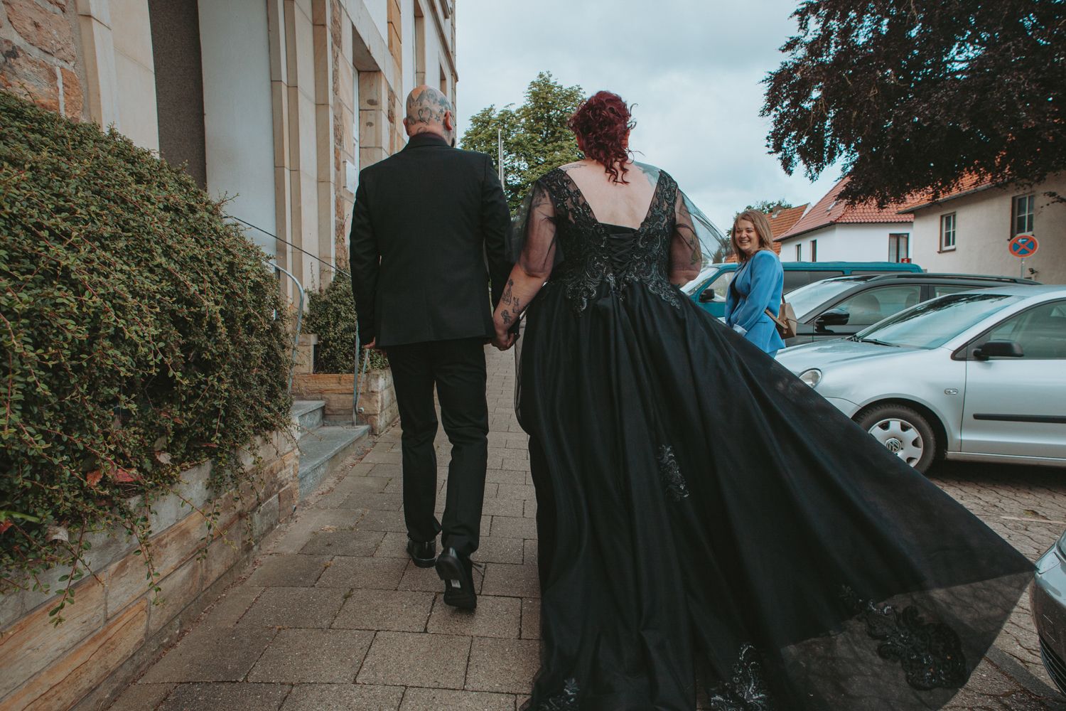 A couple walks away from camera down a sidewalk, with the person on the right wearing a flowing black formal gown.