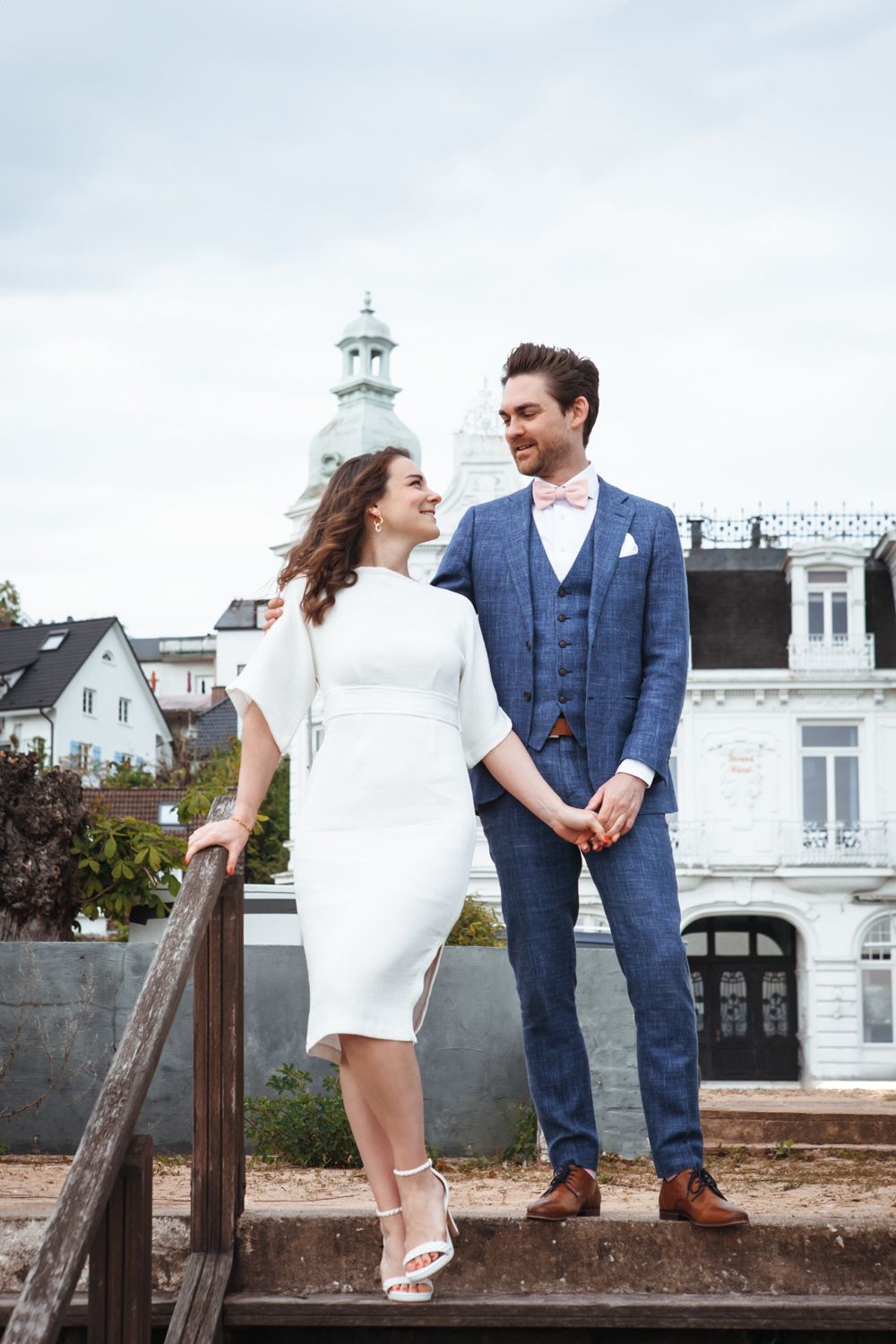 A couple in classic wedding attire stands together on stone steps against a backdrop of white buildings and a church steeple.
