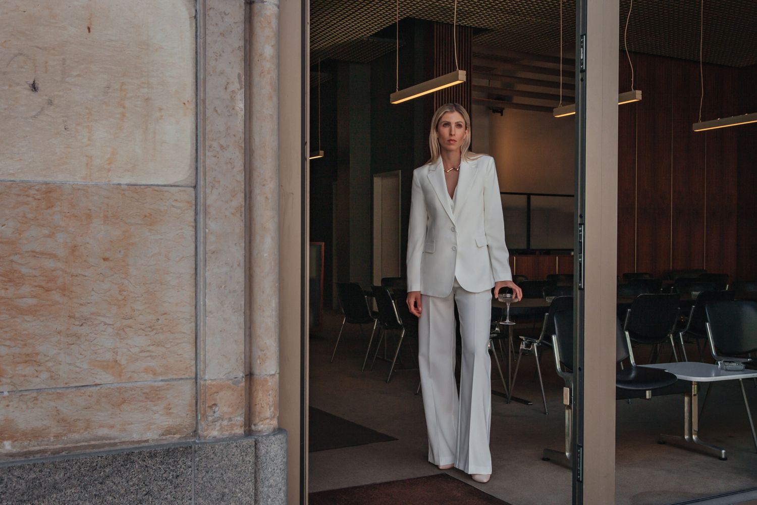 Professional wearing a stylish white pantsuit stands in a doorway of an office building with vintage chairs visible inside.