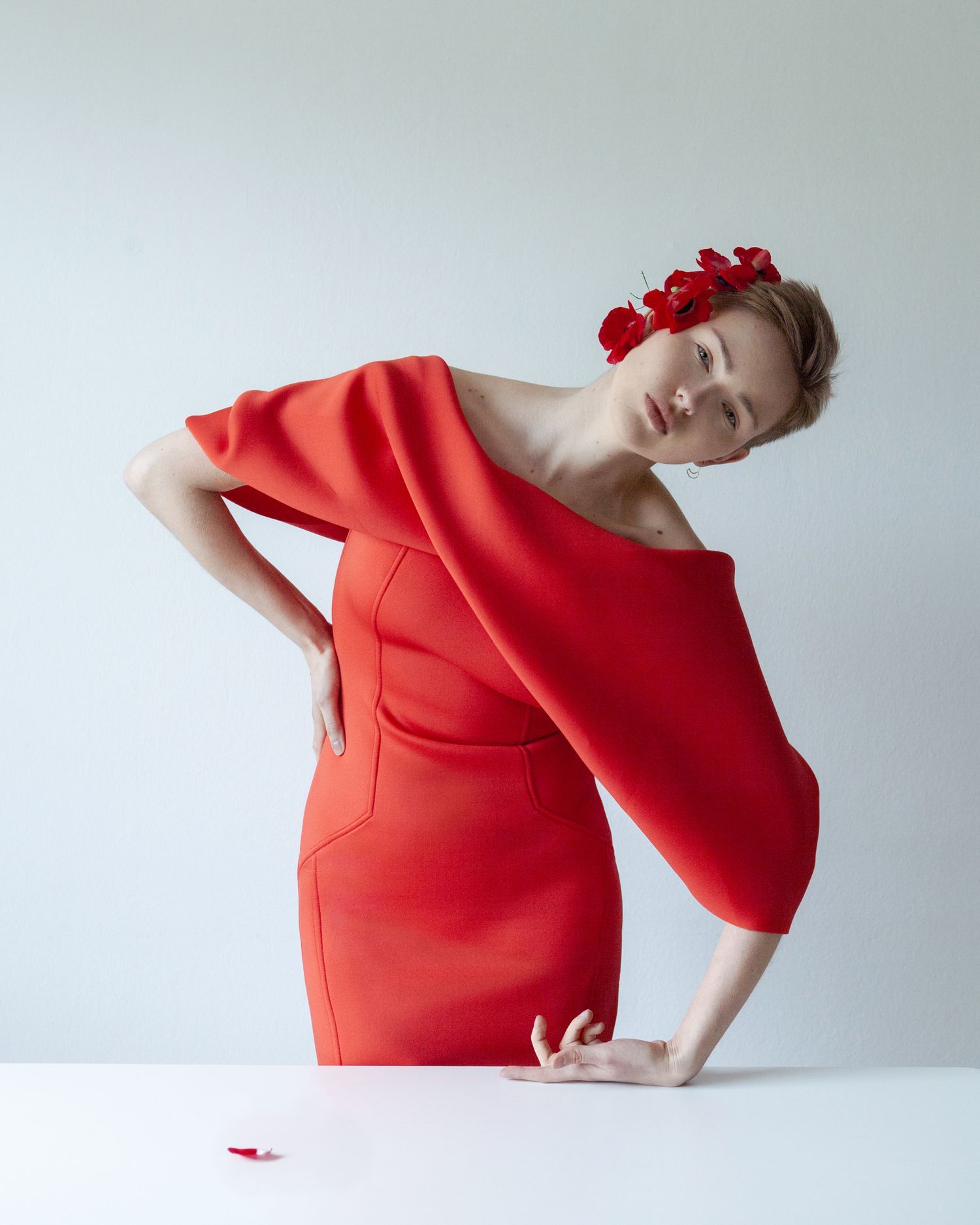 Artistic pose in flowing red dress with floral crown against white backdrop.