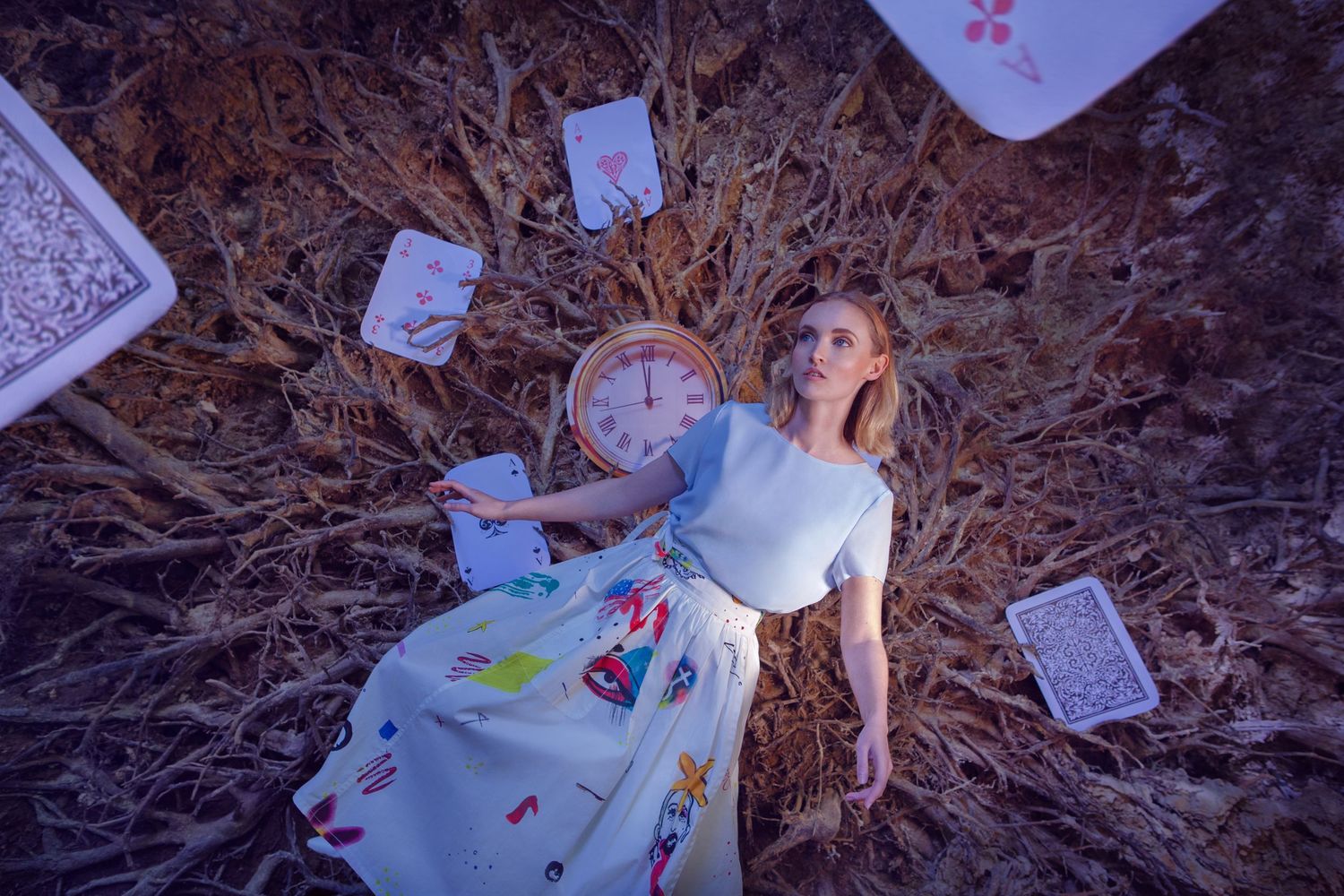 Overhead shot of a figure in a blue dress surrounded by scattered playing cards on wooden ground.