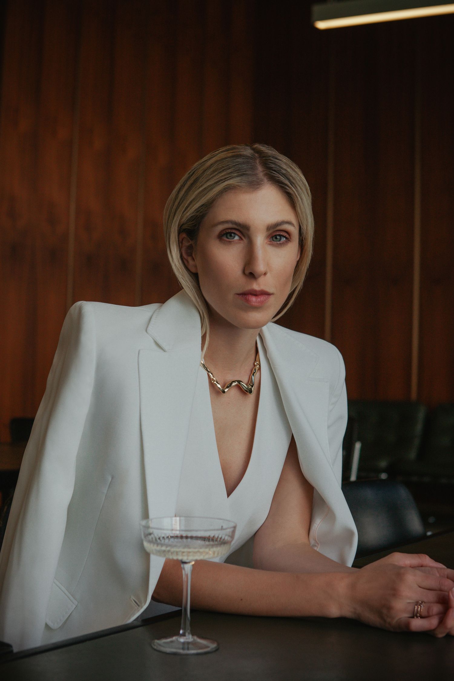Professional woman in white blazer seated at bar with cocktail glass and statement necklace in moody lighting.