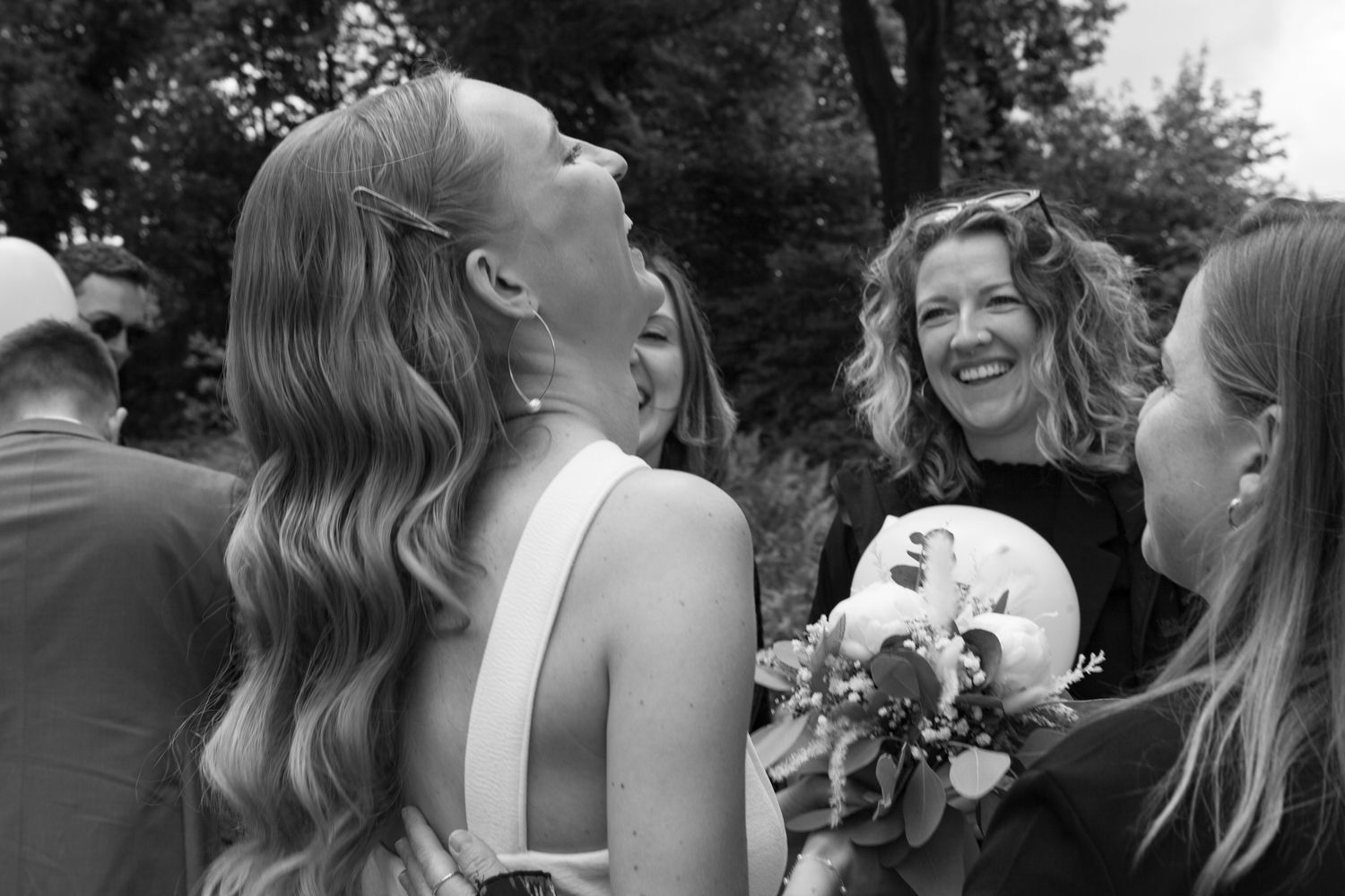Black and white candid photo showing people sharing a joyful moment of laughter at an outdoor celebration with a bouquet.