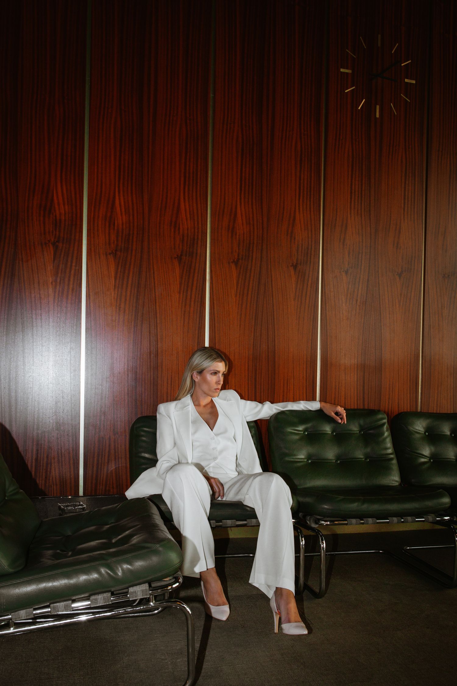Woman in white suit sitting on vintage green leather couch against wood paneled wall.