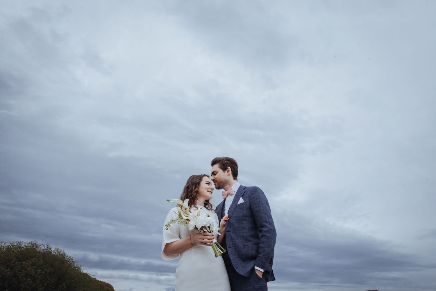A couple in wedding attire sharing a romantic moment against a dramatic cloudy sky backdrop.