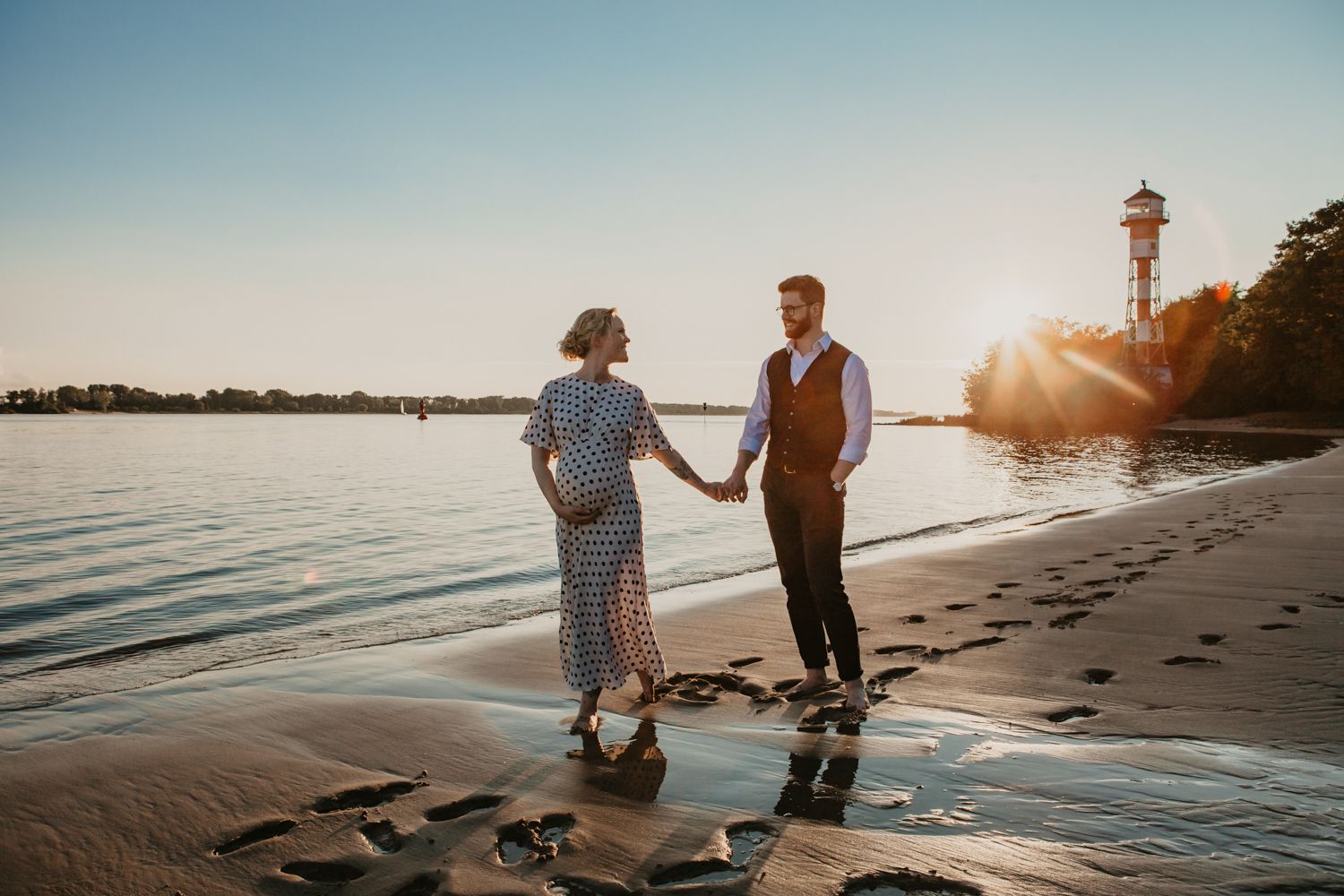 A couple strolls along the shoreline at sunset with a lighthouse visible in the distance.
