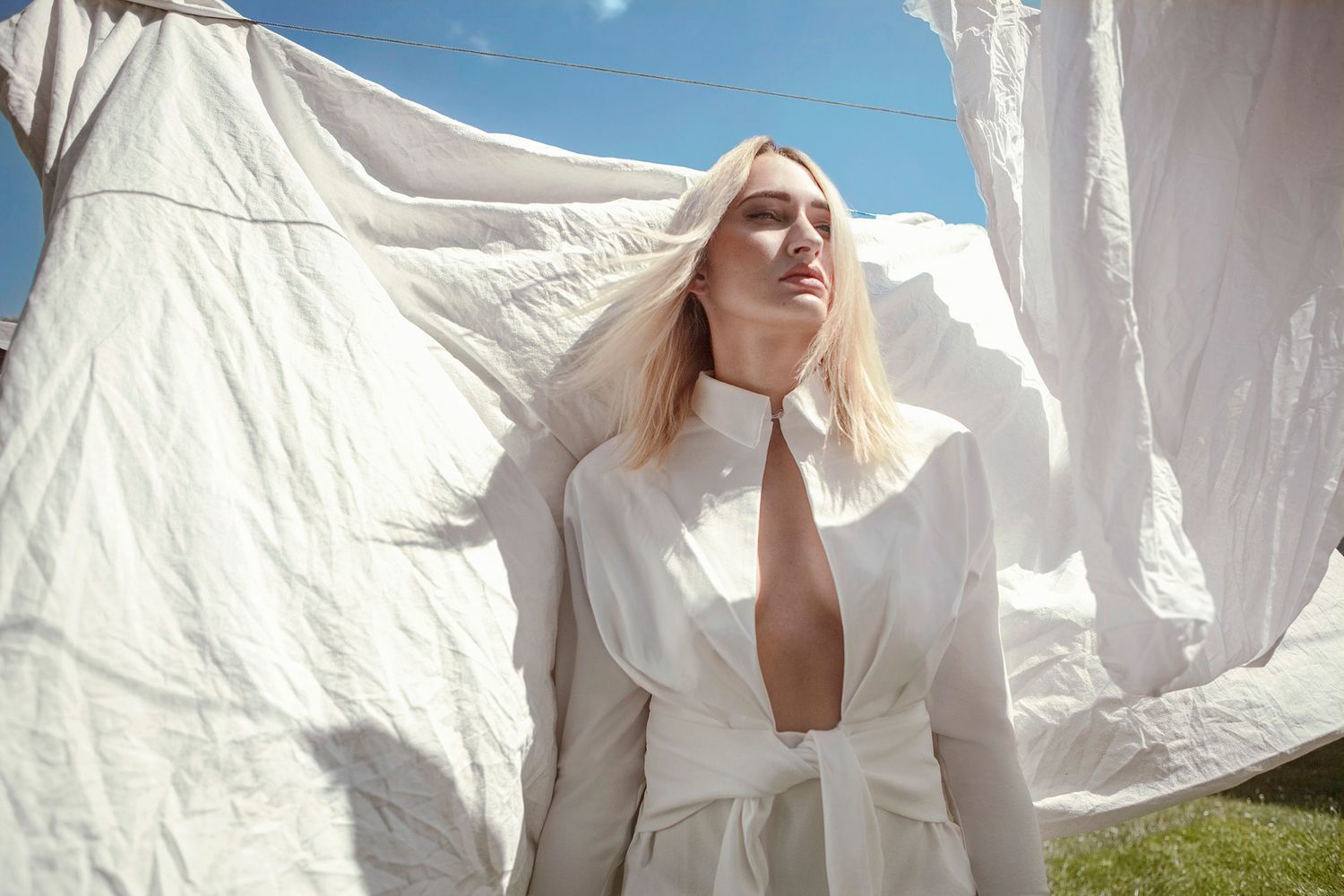 Fashion model in white draped garment poses against billowing white sheets outdoors.