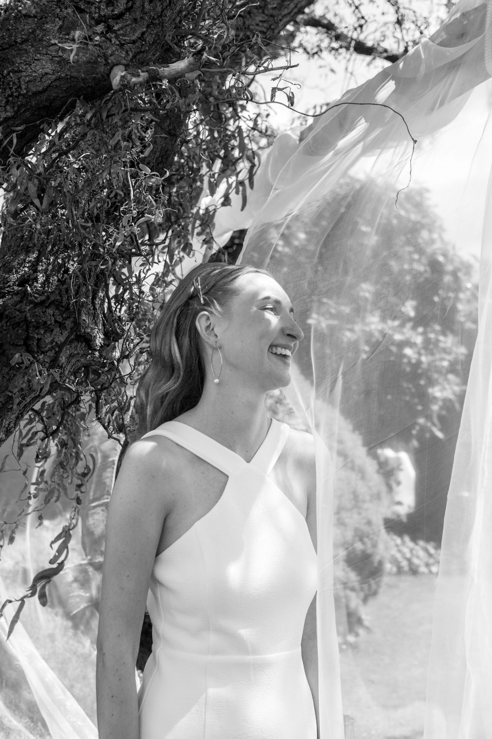 Black and white photo of a bride in a halter neck wedding dress smiling under hanging vines outdoors.