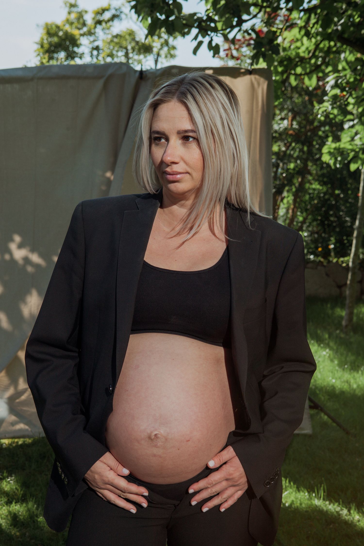 An outdoor maternity photo showing a pregnant figure in a black blazer and crop top against a garden backdrop.