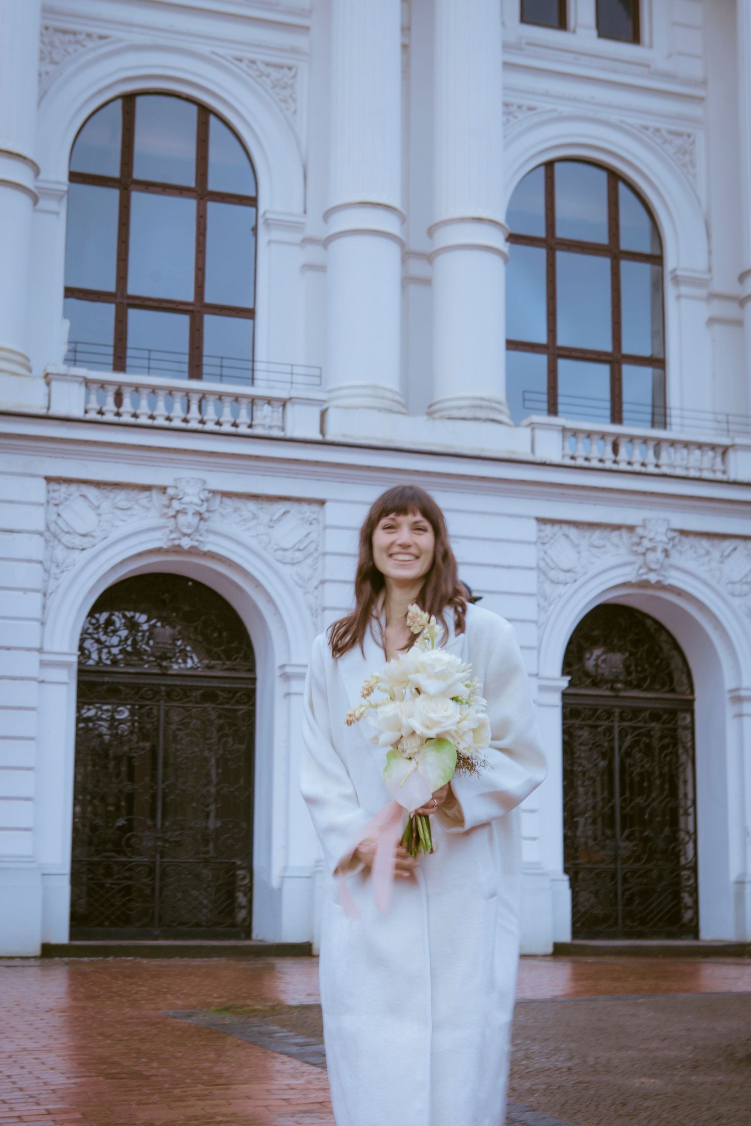 Bride in modern white outfit holding bouquet poses in front of classical architecture building.