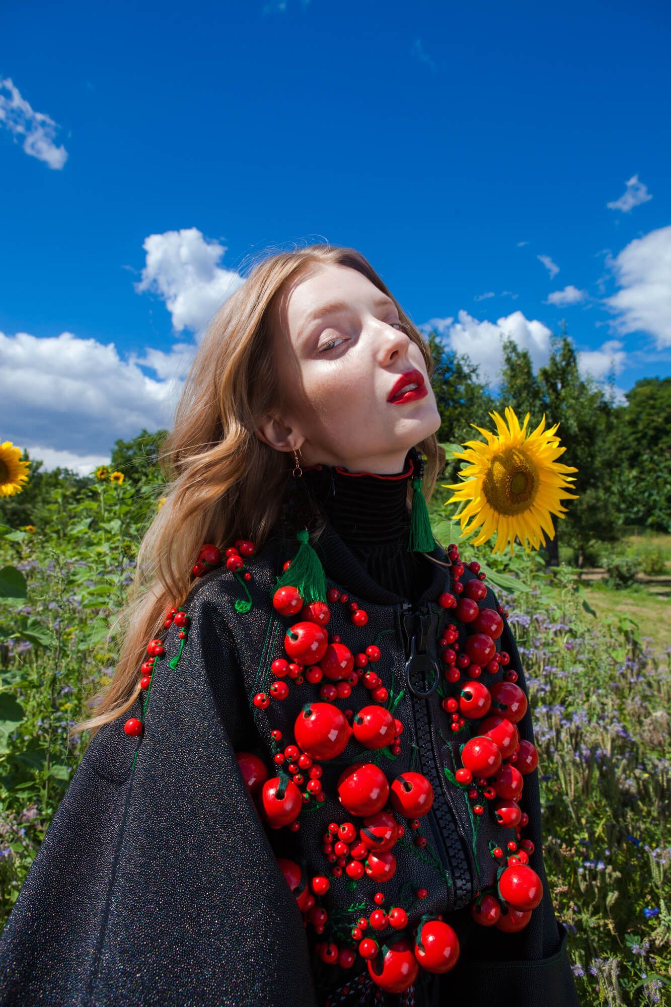 Artistic fashion portrait in a sunflower field featuring a black dress with red floral details against bright blue sky.