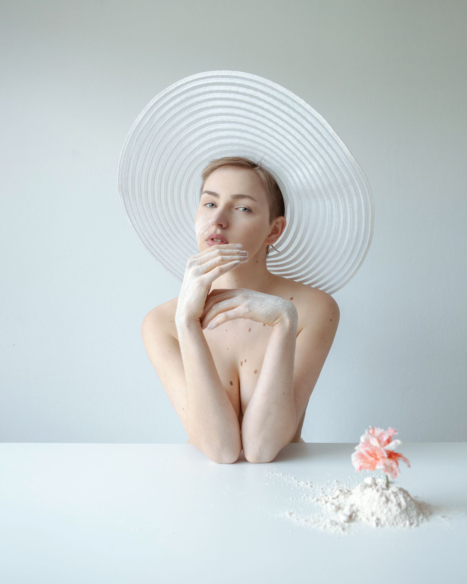 Minimalist portrait with large white sun hat and delicate pink flower on table.