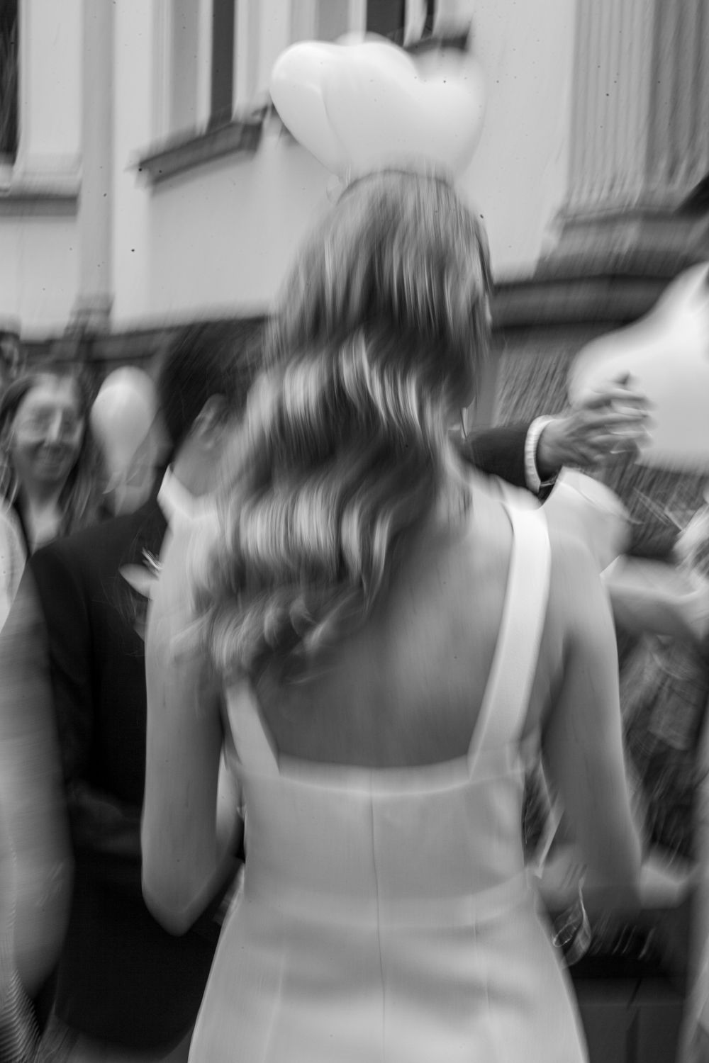 Black and white photo of someone in a white dress with long wavy hair holding heart shaped balloons at an event.