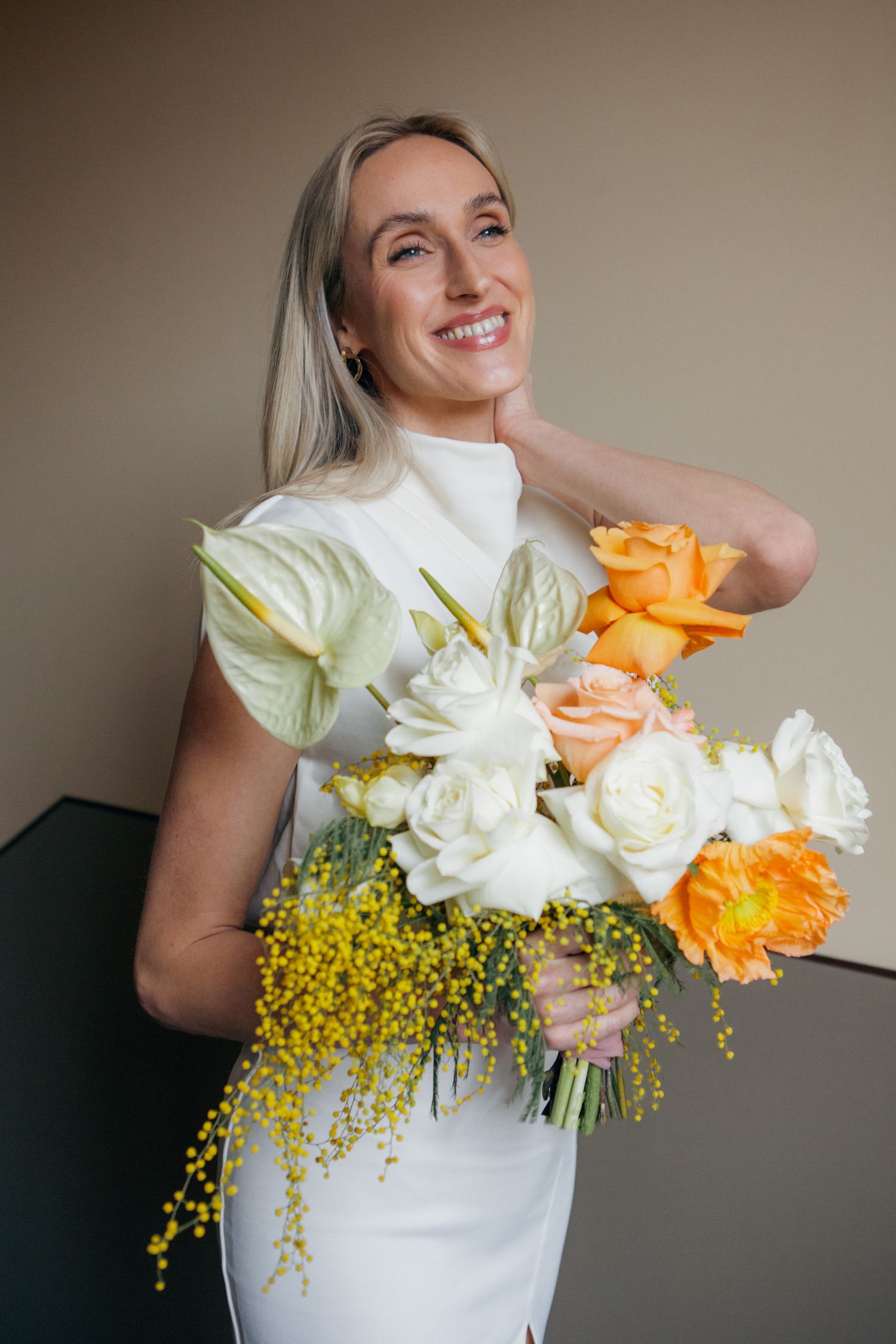 Woman in white holding vibrant bouquet with roses, orange flowers and yellow mimosa blooms against beige wall.