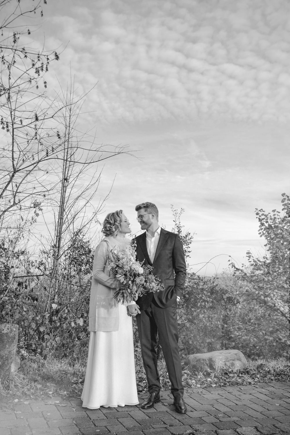 A romantic black and white photo of a couple sharing an intimate moment outdoors on a brick pathway surrounded by nature.