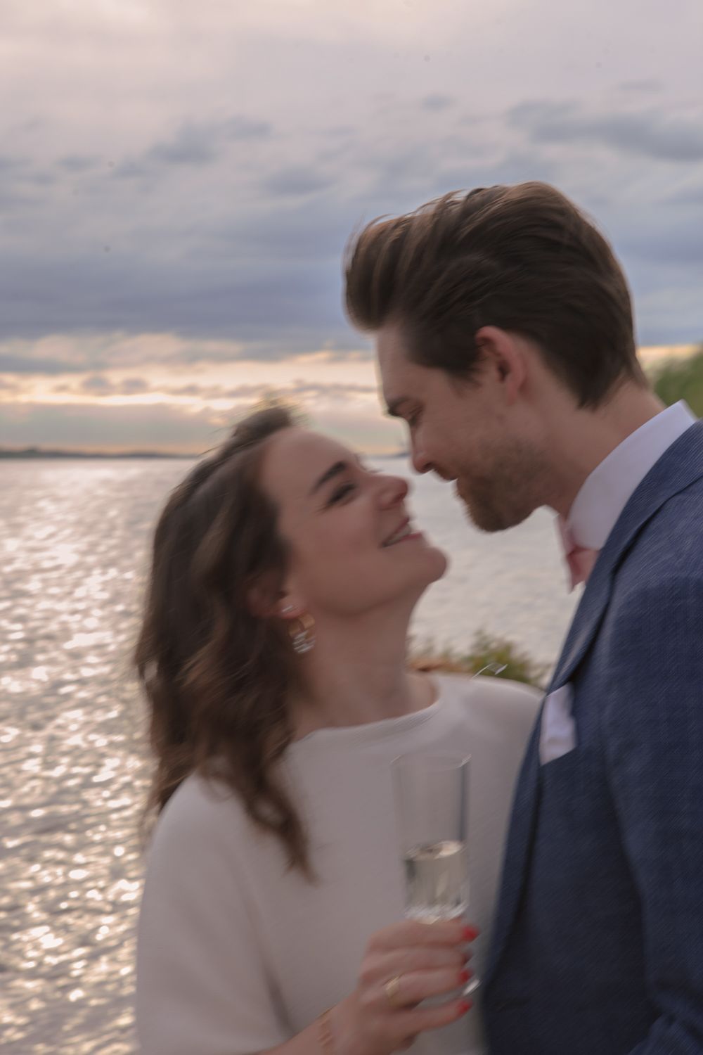 Romantic couple sharing an intimate moment at sunset by the ocean while holding champagne glasses.