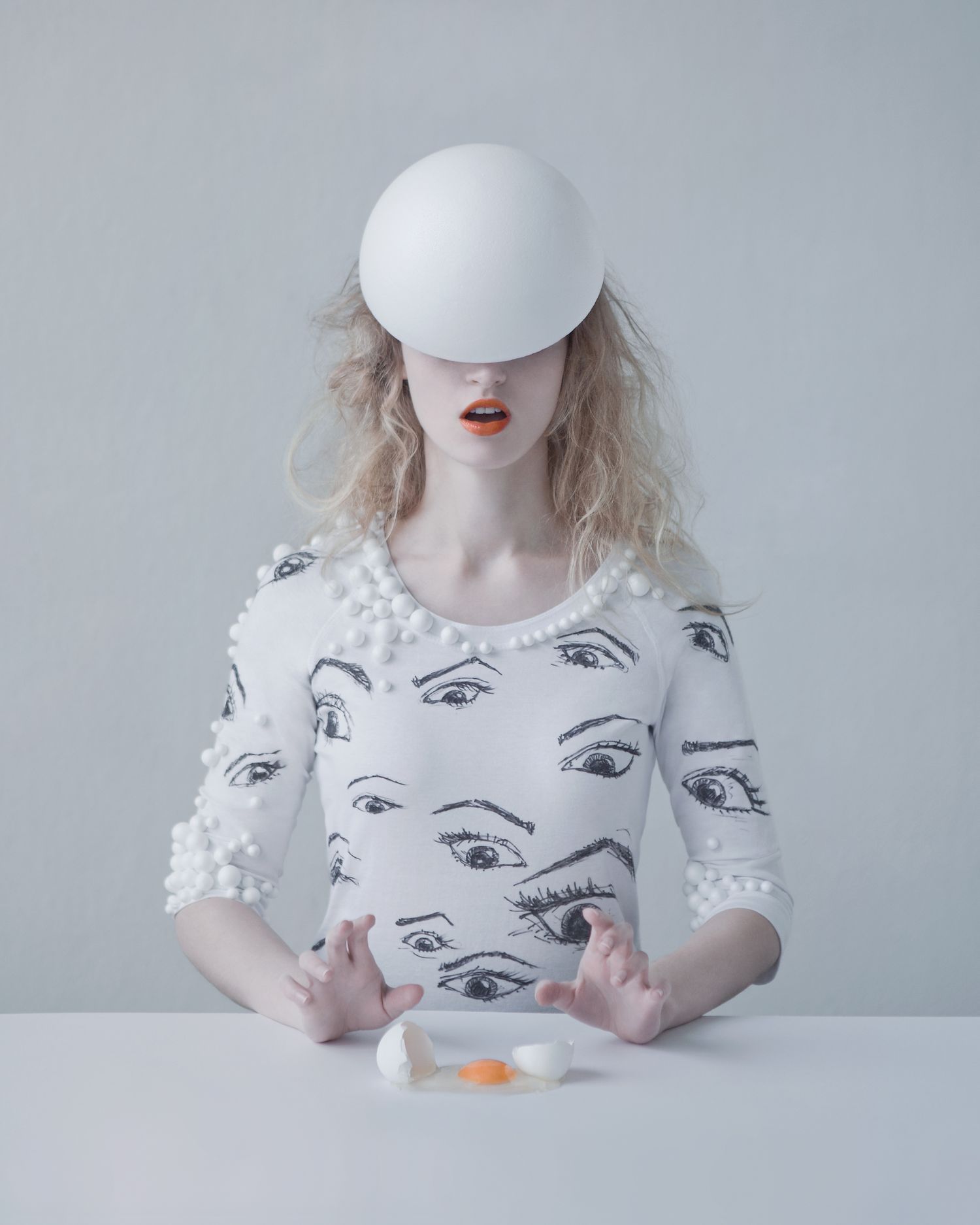 Surreal portrait of someone wearing white top with eye pattern and white sphere covering their head at a minimalist table setting.