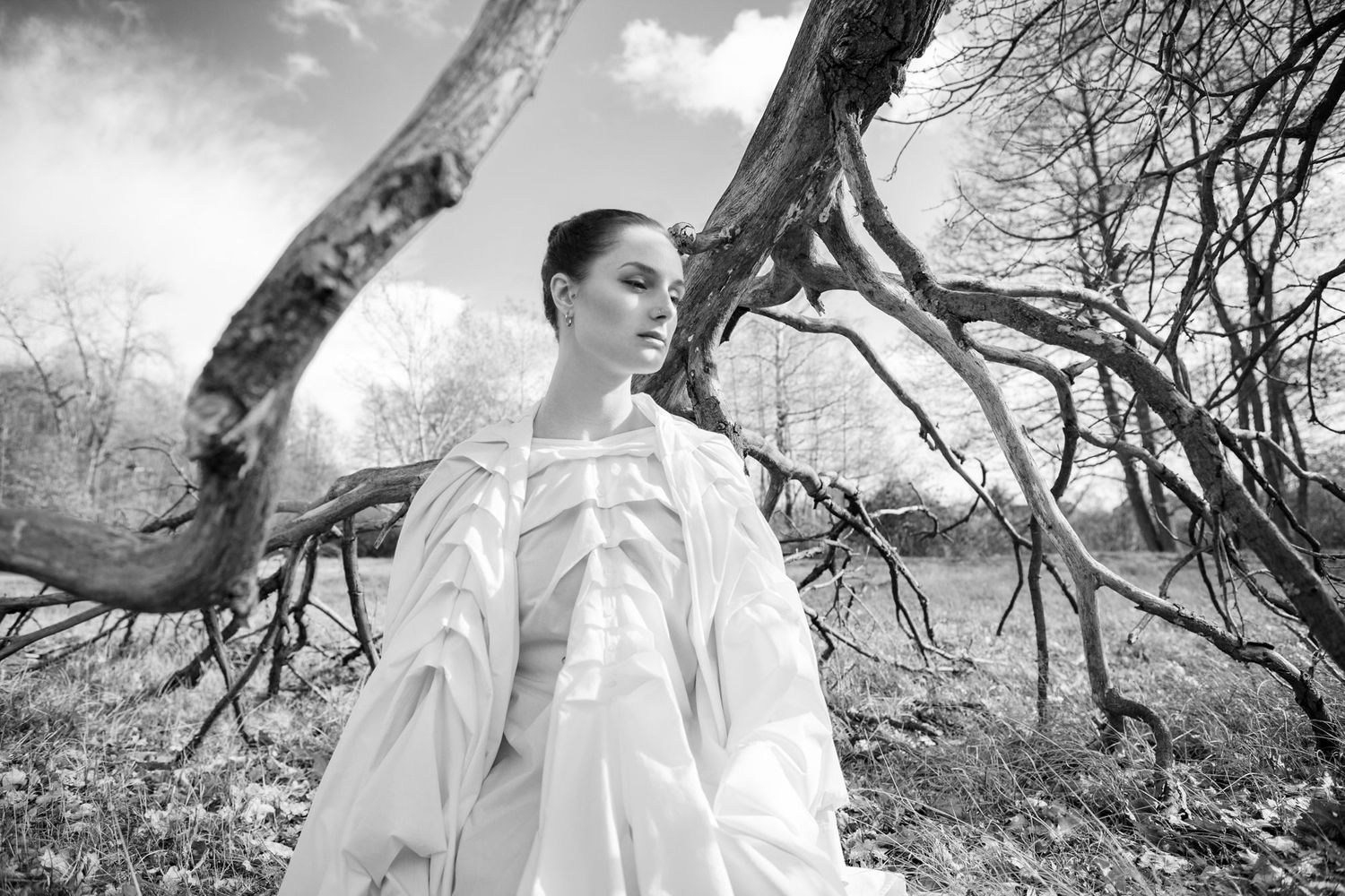 Fashion model wearing flowing white garments stands against dramatic winter tree branches.