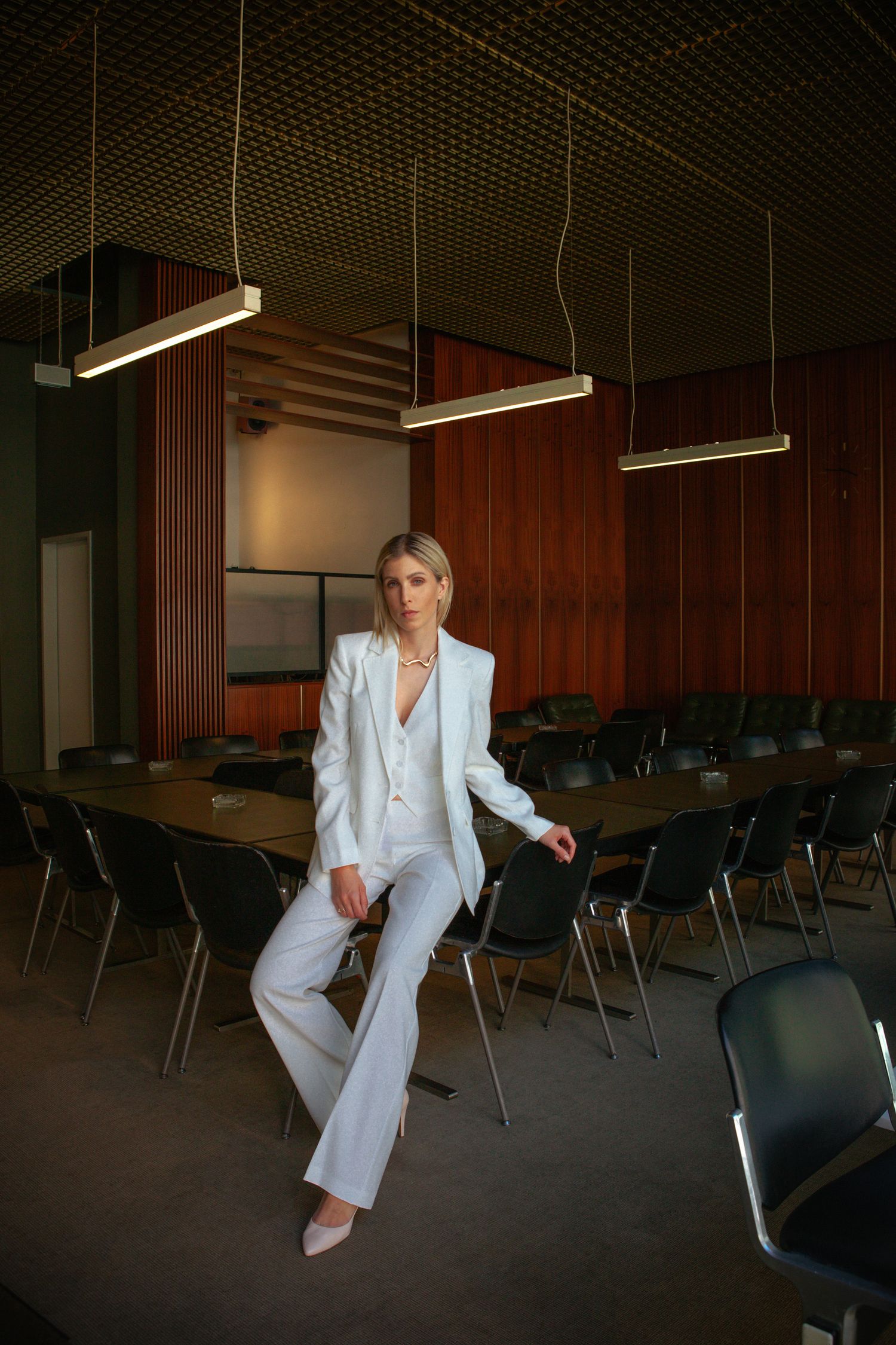 Stylish woman in all-white suit poses in modern conference room with hanging lights.