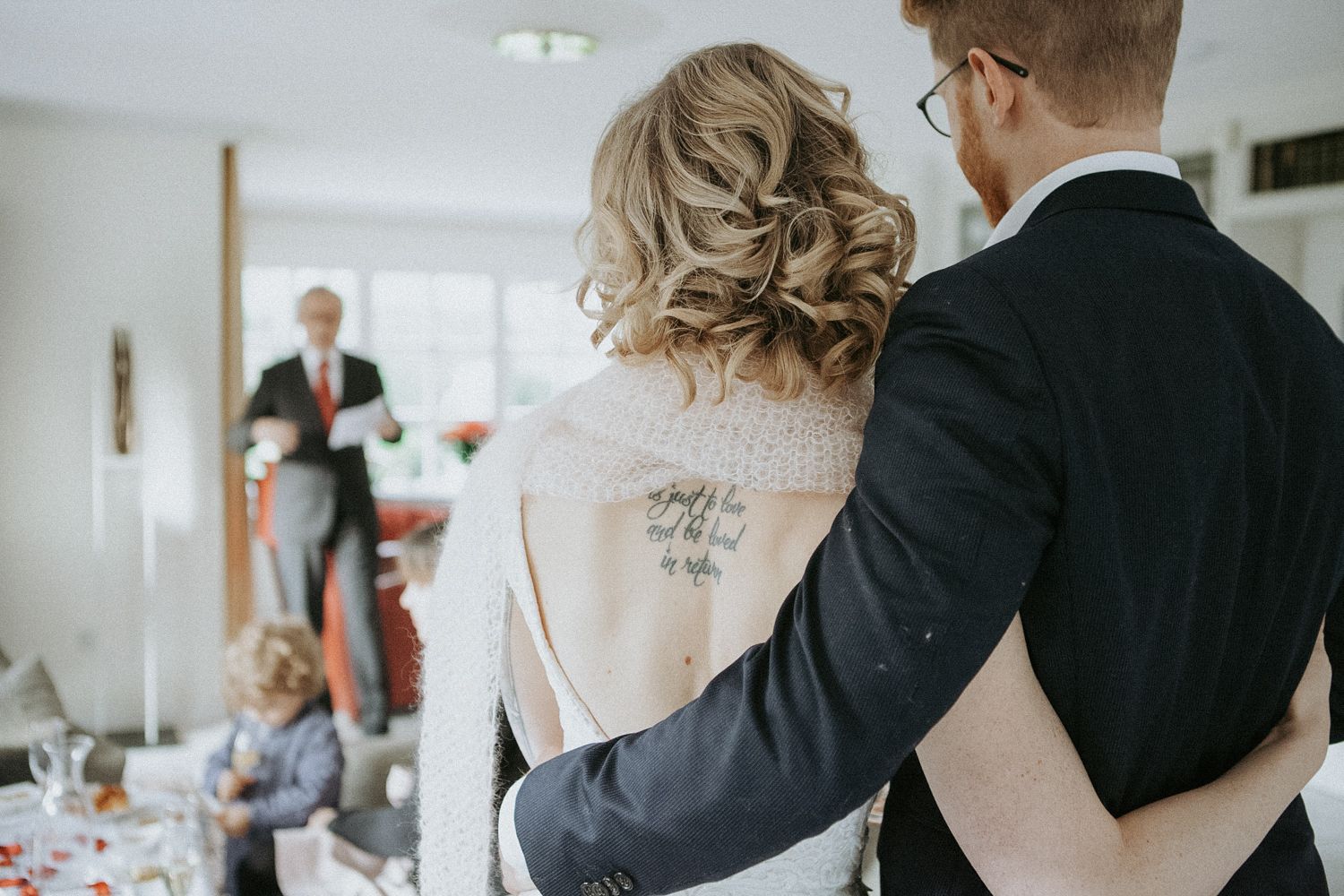 A tender wedding moment captured from behind as people gather at a reception in a bright indoor venue.