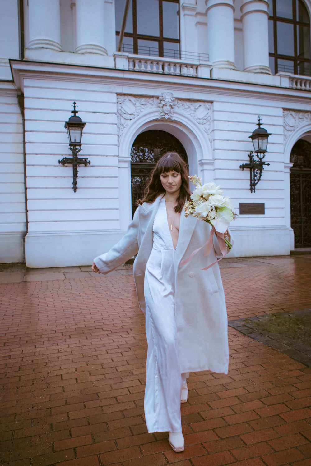 A model in a flowing white shirt dress walks gracefully across terracotta tiles outside an elegant white building.
