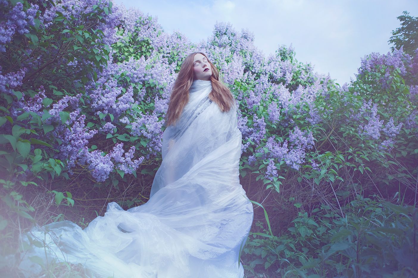 Ethereal figure in flowing white dress poses dramatically among blooming purple lilac bushes in dreamy garden setting.