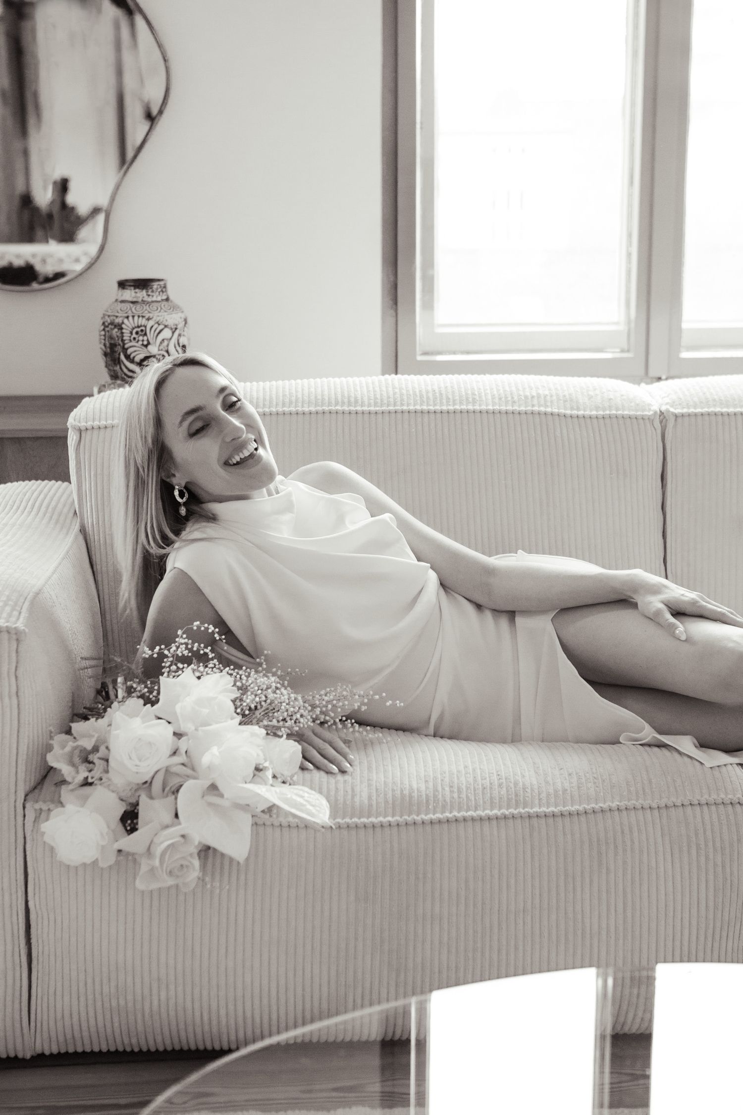 Black and white photo of a bride relaxing on a white couch with flowers.