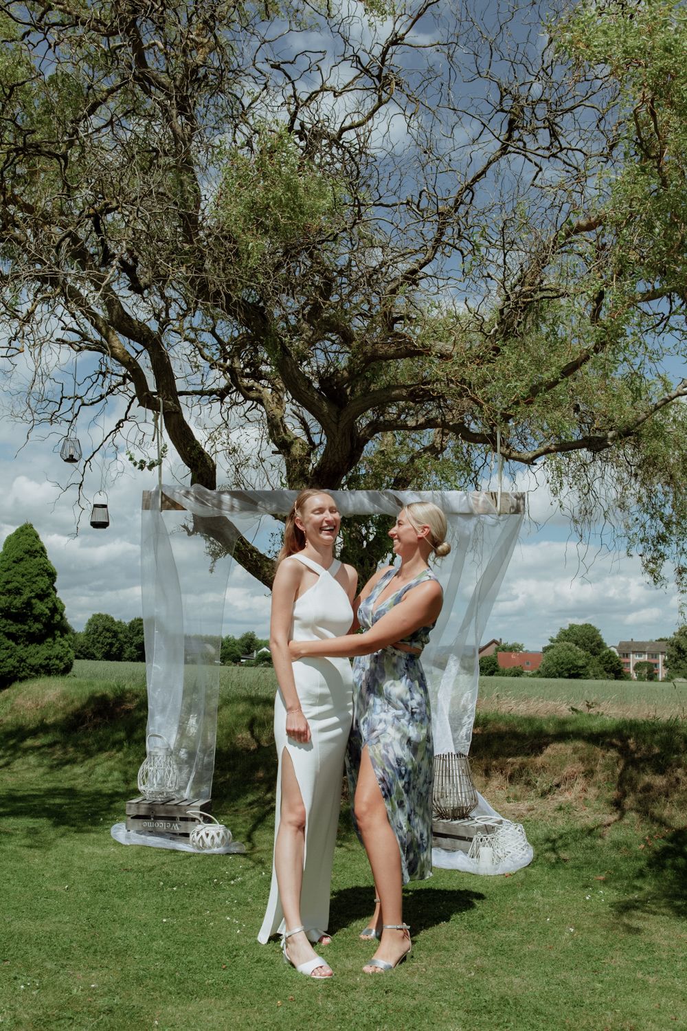 Two people dressed in white share intimate moments in a grassy outdoor setting with hanging glass candle holders in trees.