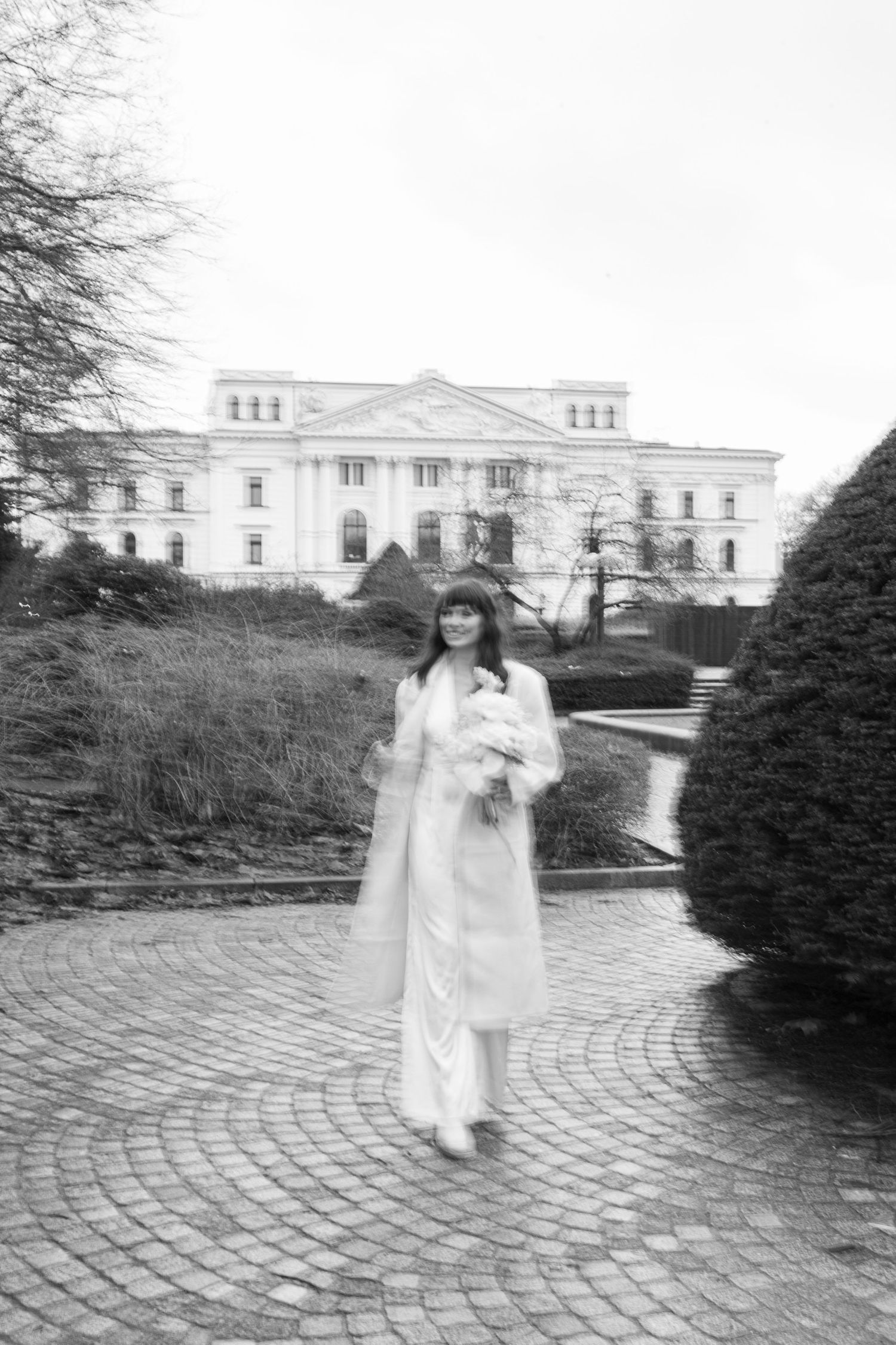 Black and white photo of figure in white coat standing before grand historic mansion.