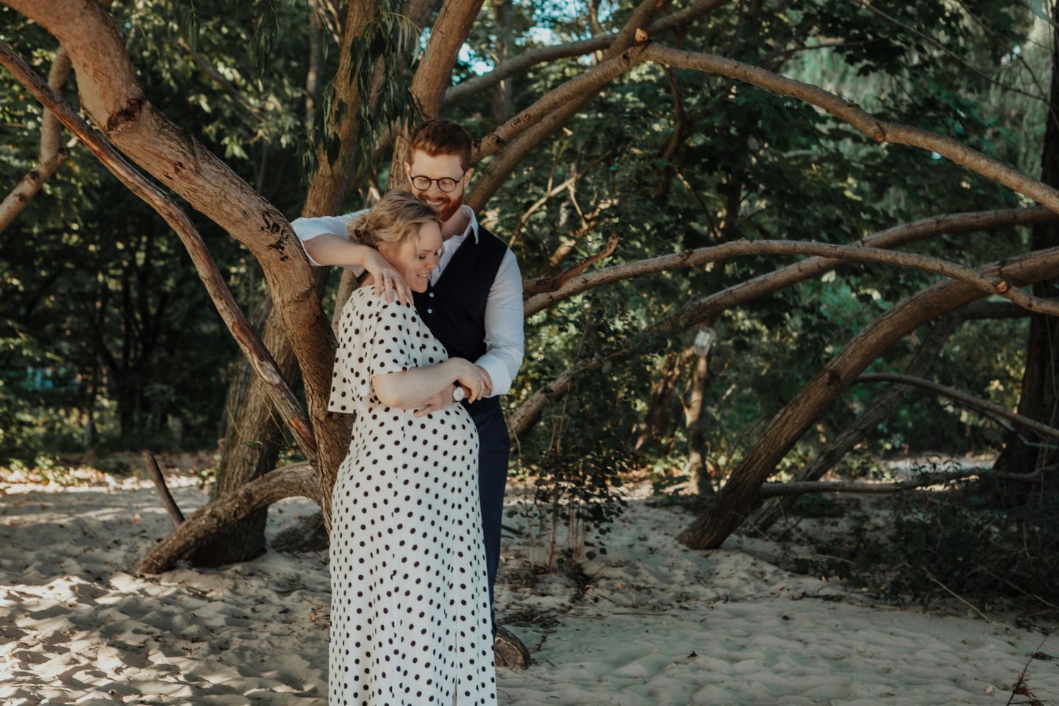 A couple shares an intimate moment under a large sprawling tree on a beach.