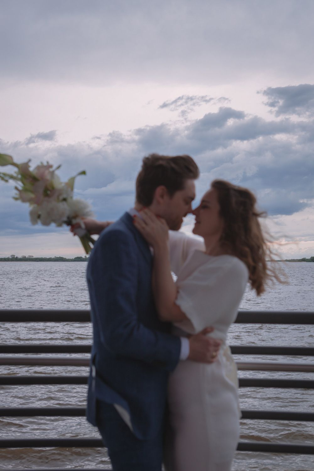 A romantic couple embraces on a waterfront pier at sunset with flowers and cloudy skies in the background.
