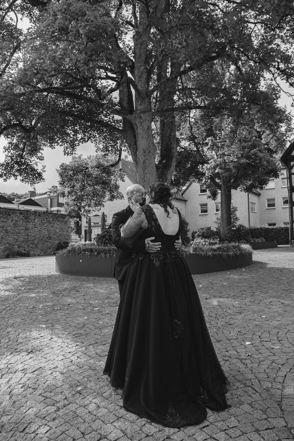 A figure in a flowing black dress and leatherwork stands on a curved stone pathway in a garden setting.