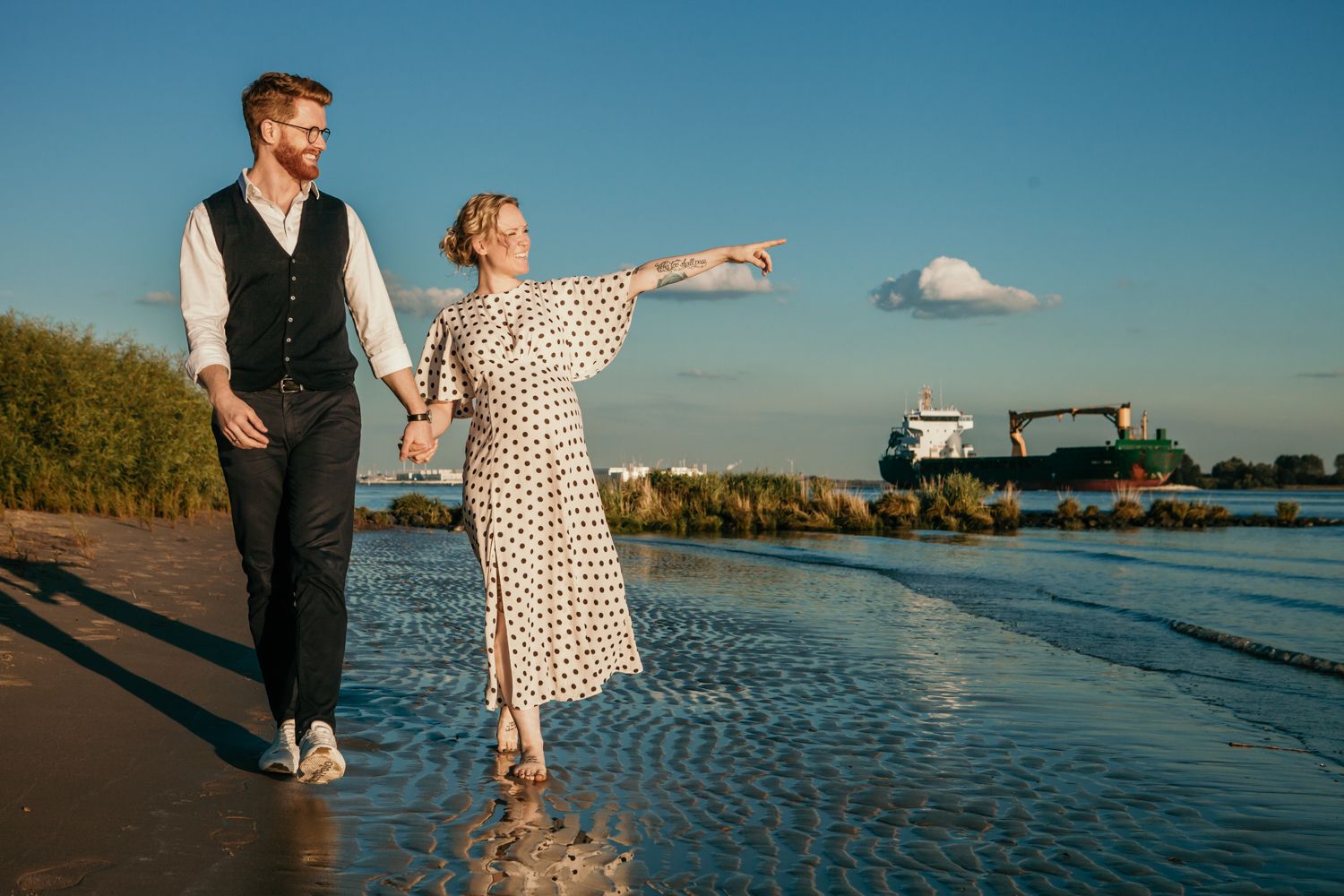 A couple in vintage style clothing walks along a beach at sunset with a lighthouse in the background.